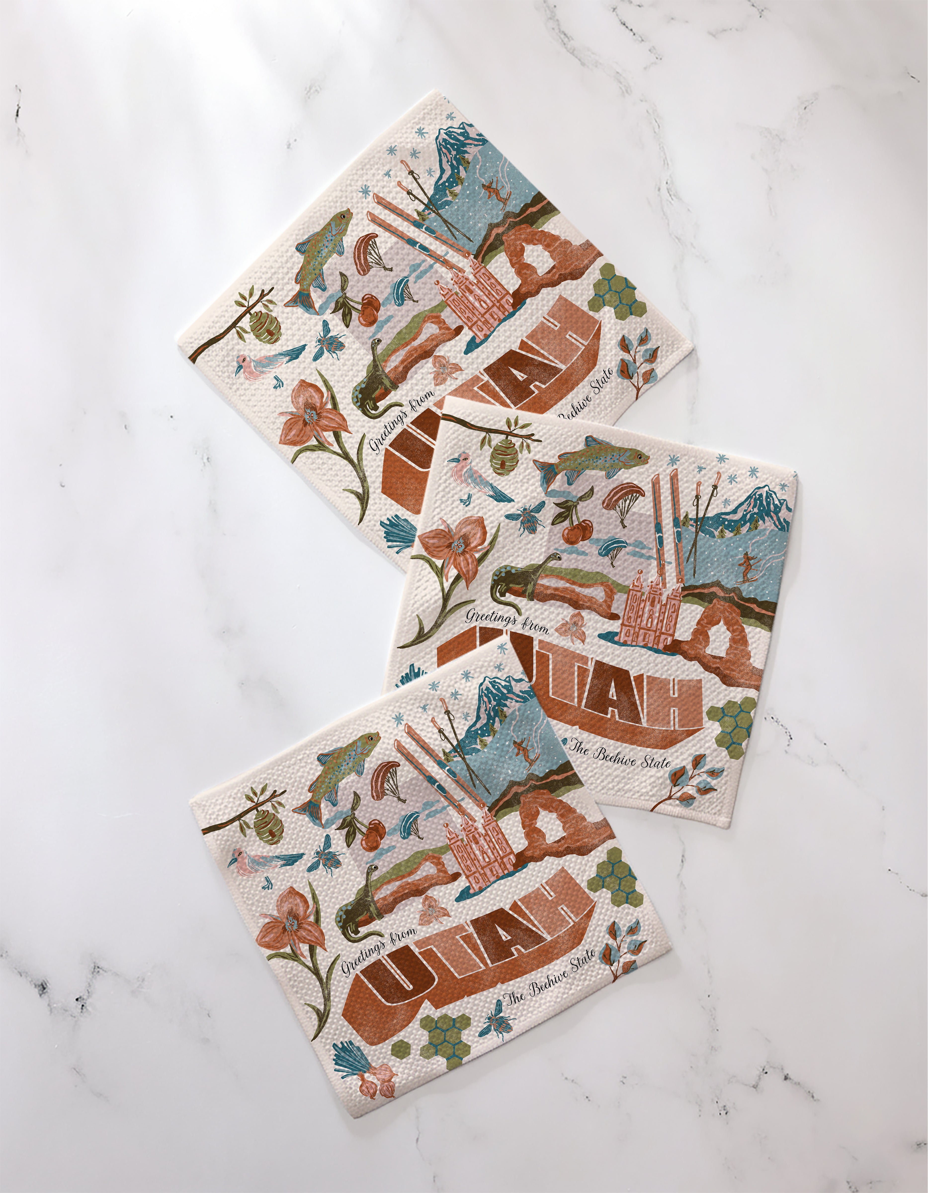 Three "Greetings From Utah" napkins by Geometry, featuring mountain, flower, and landmark illustrations in brown, blue, and green on recycled fabric, are arranged on a white marble surface.