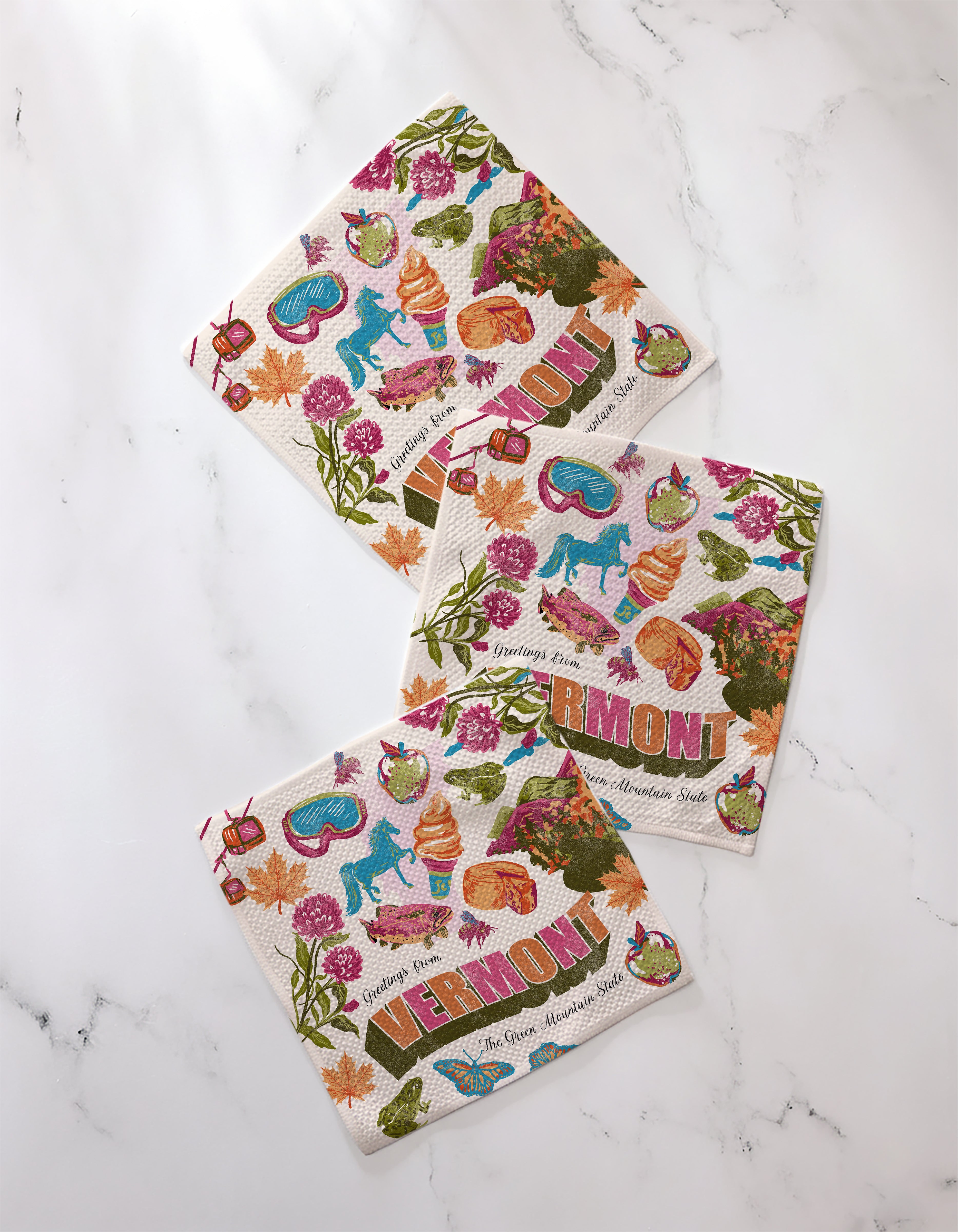 Three "Greetings From Vermont" paper napkins by Geometry, featuring colorful Vermont-themed illustrations, are displayed on white marble next to a recycled dishcloth set.