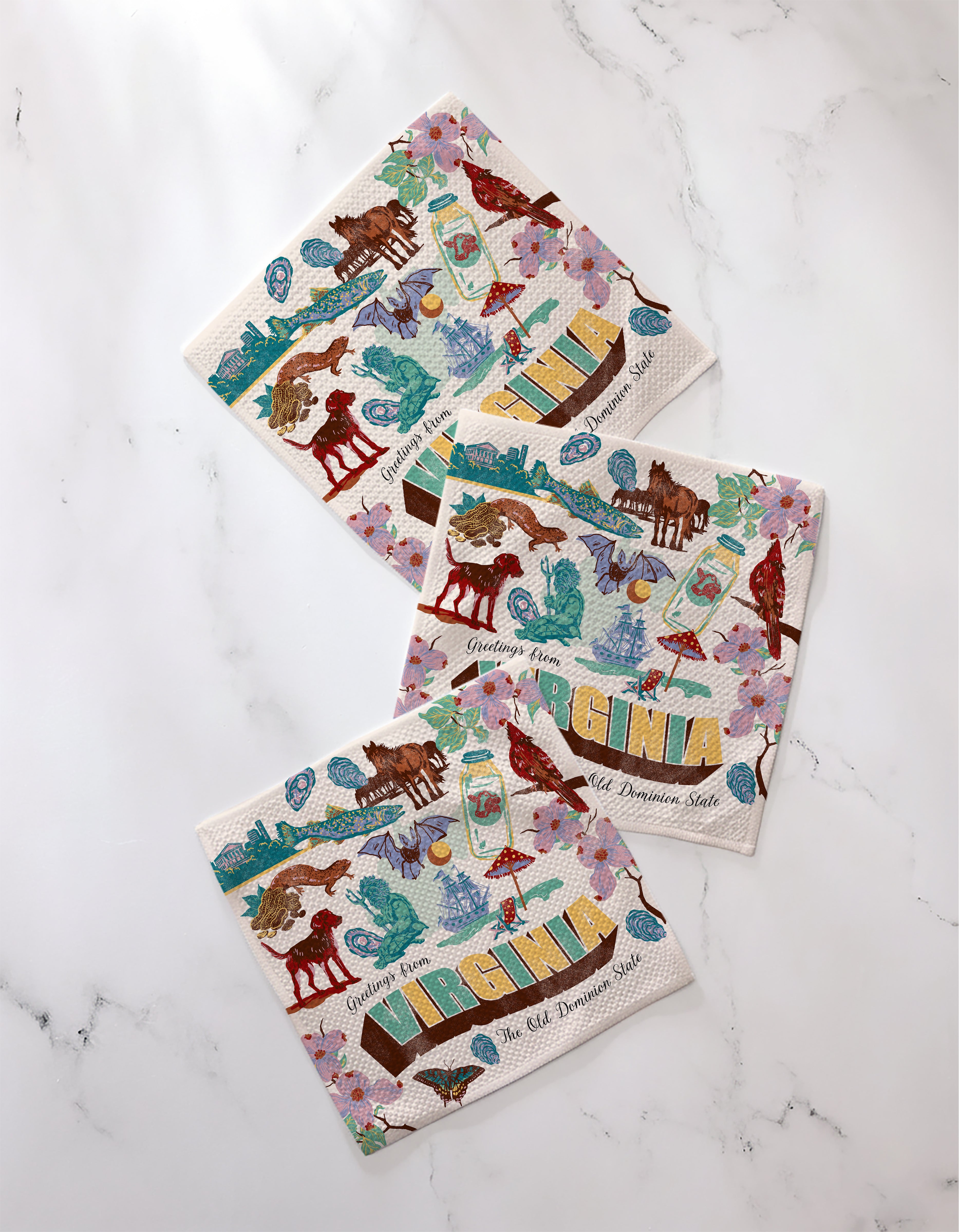 Three "Greetings From Virginia" paper napkins by Geometry, featuring colorful horses, flowers, and state icons with bold VIRGINIA lettering, are arranged on white marble. Inspired by absorbent kitchen towels.