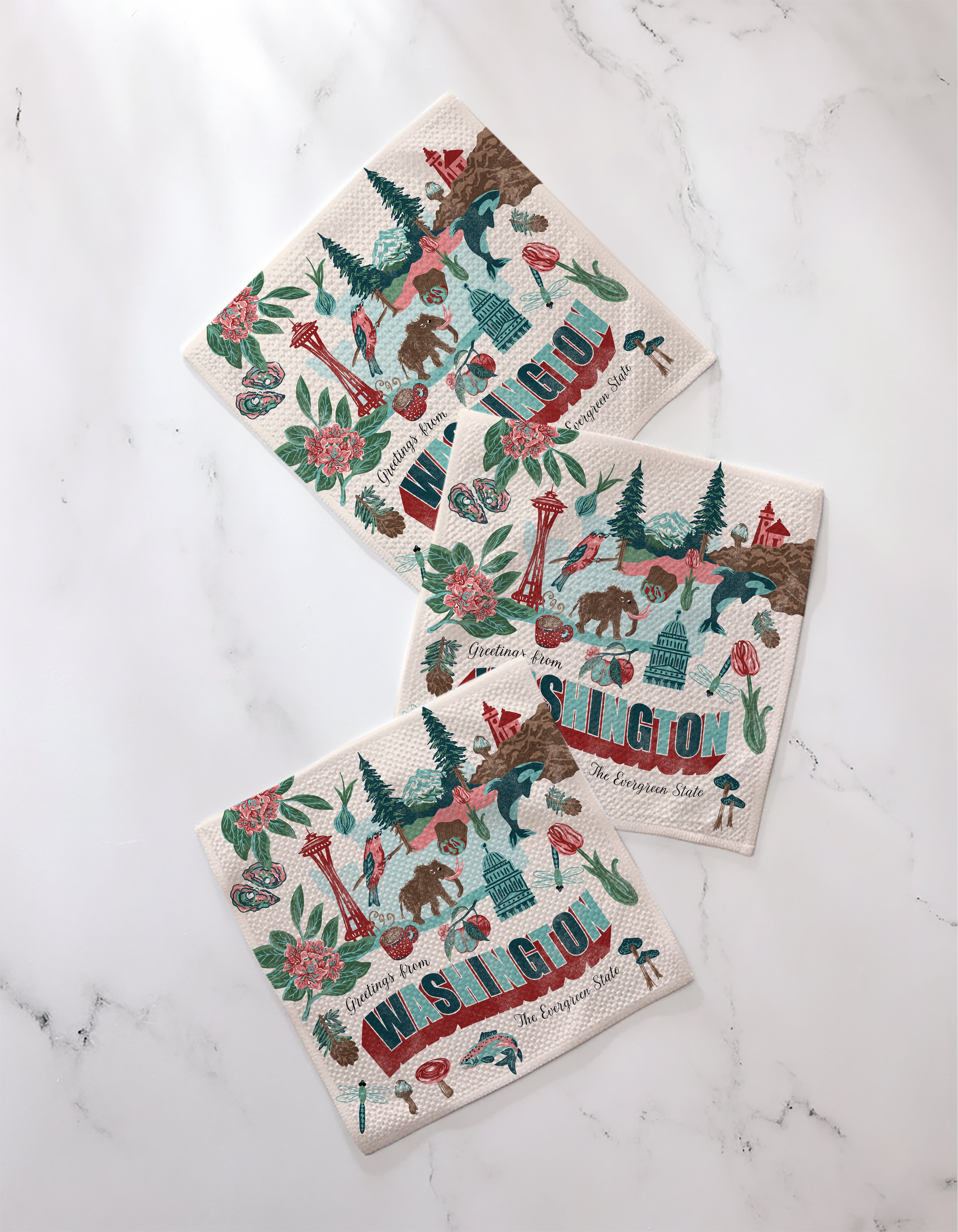 Three "Greetings From Washington" paper napkins by Geometry, made from recycled materials and decorated with colorful Washington state symbols, are displayed on a white marble surface.