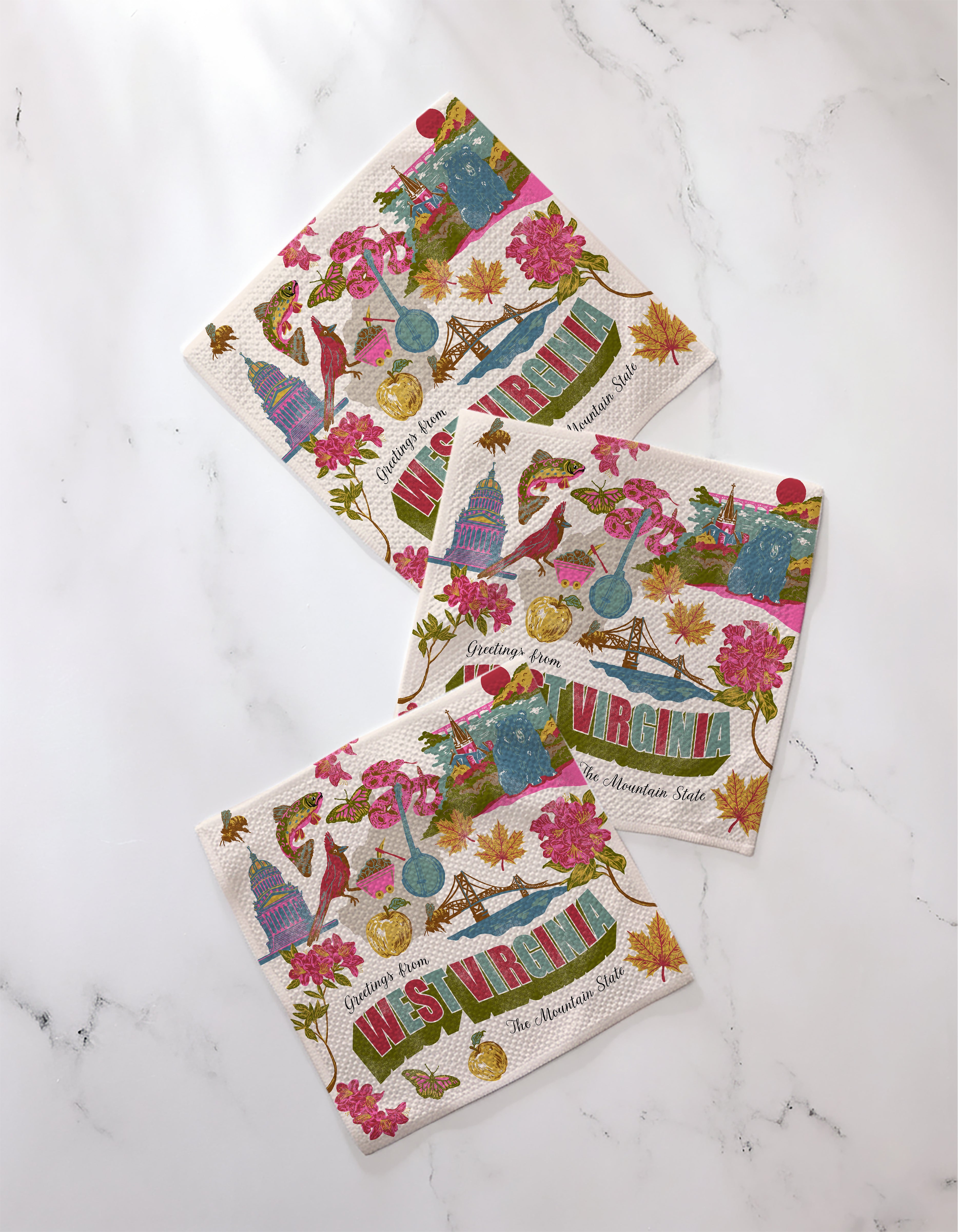 Three colorful "Greetings From West Virginia" paper napkins by Geometry, made from recycled materials, feature state-themed illustrations—flowers, a bluebird, mountains, apples, and landmarks—arranged on a white marble surface.