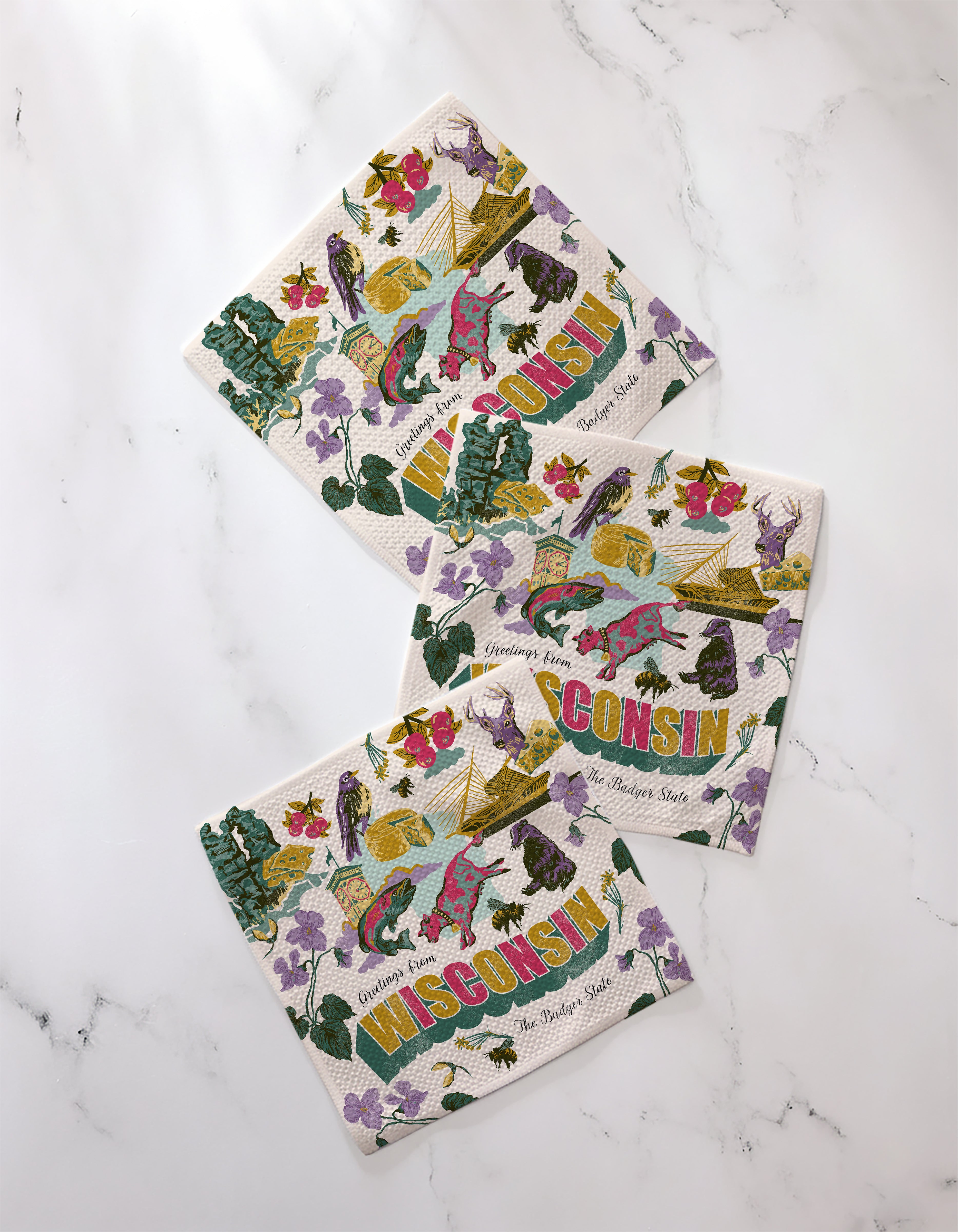 Three "Greetings From Wisconsin" napkins by Geometry feature colorful state-themed illustrations and the word WISCONSIN, with flowers, birds, and symbols on recycled materials, displayed on a white marble surface.
