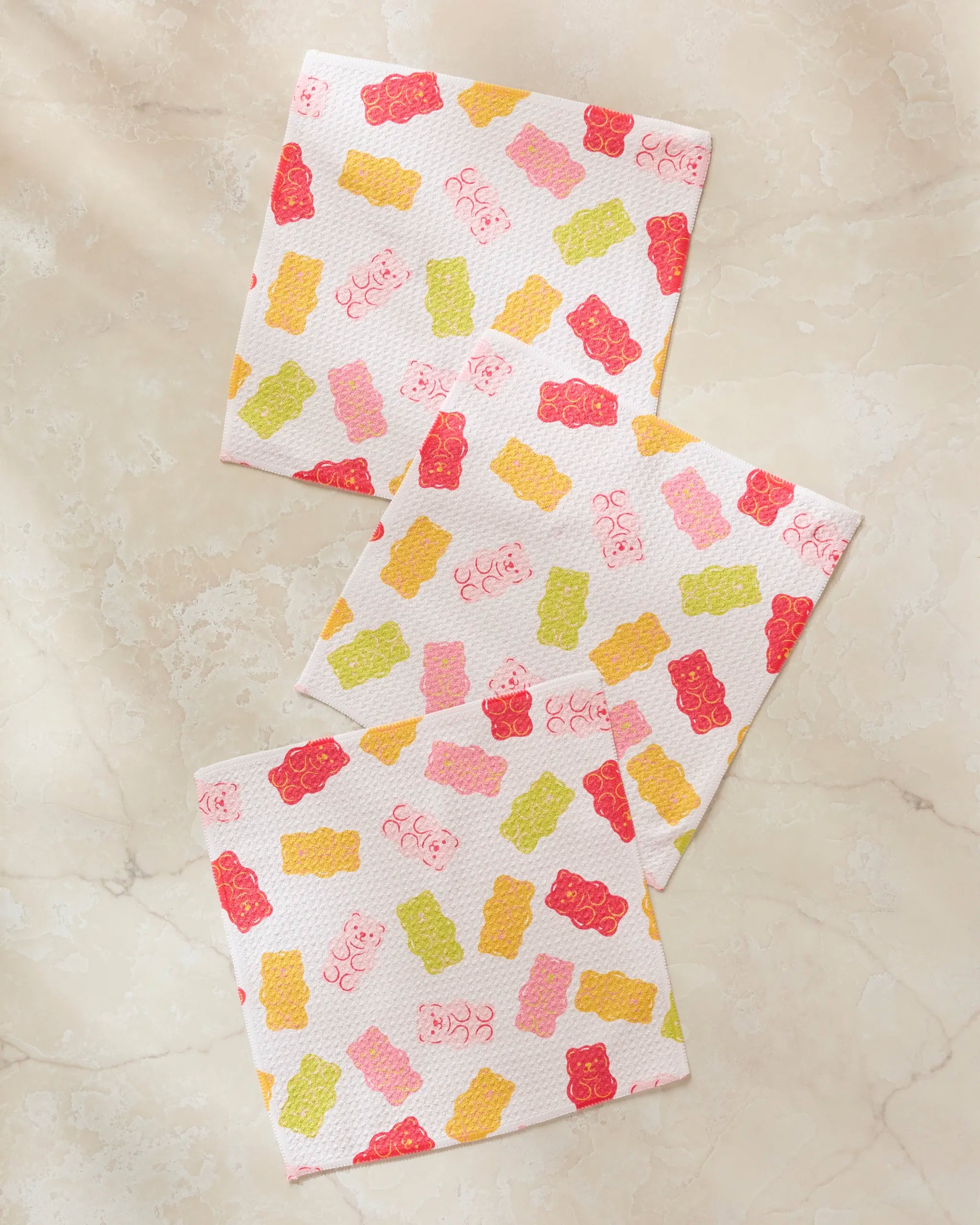 Three Gummy Bear paper napkins by Vaishali Design Studio, made from recycled materials and featuring colorful red, yellow, green, and pink gummy bear designs, are displayed on a light beige marble surface.