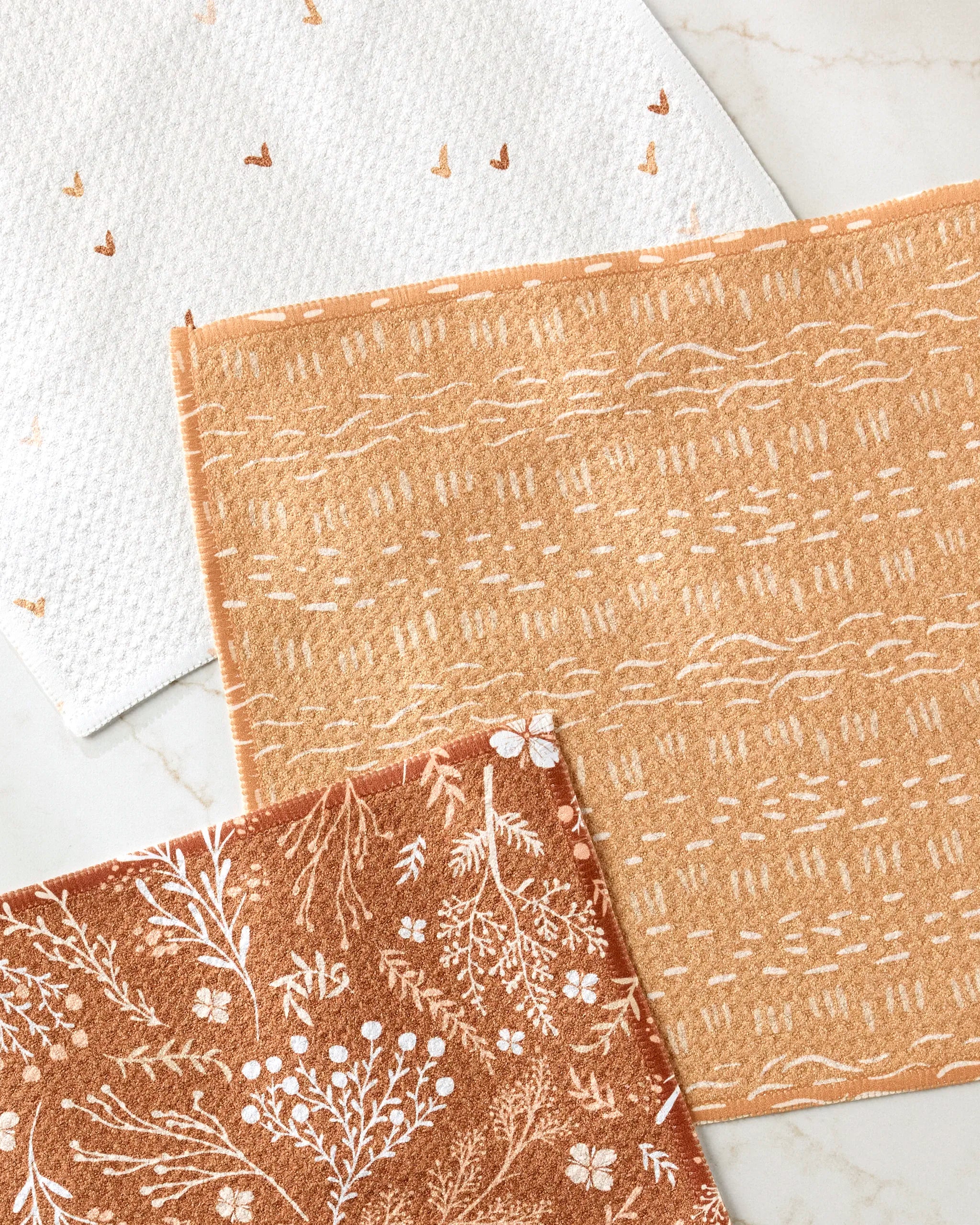 The Danelys Sidron "Gust of Wind" set includes three patterned cloths: white with brown hearts, tan with abstract lines, and rust with white florals—eco-friendly dishcloths made from recycled materials.