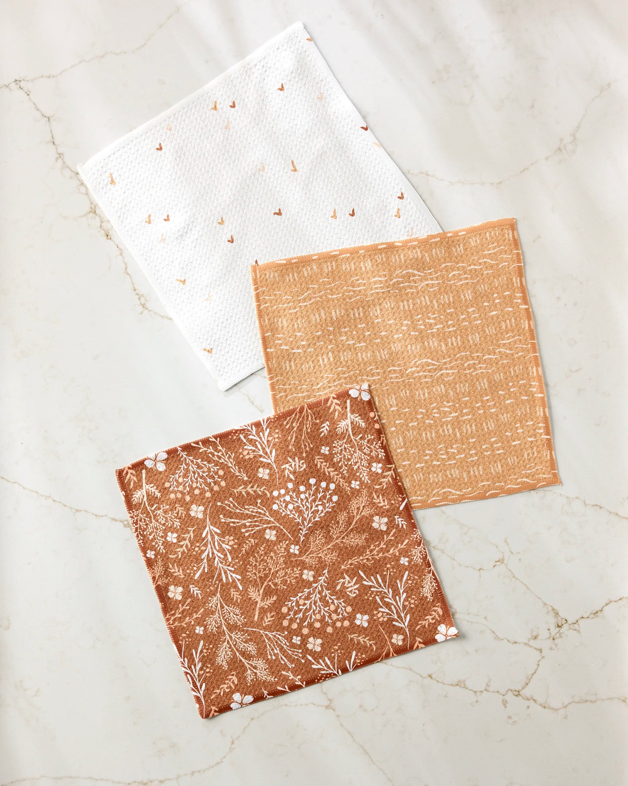 Danelys Sidron’s “Gust of Wind” set features three reusable cloth napkins: white with orange dots, terracotta with a white floral print, and one with a subtle geometric design, all made from recycled materials and displayed on light marble.