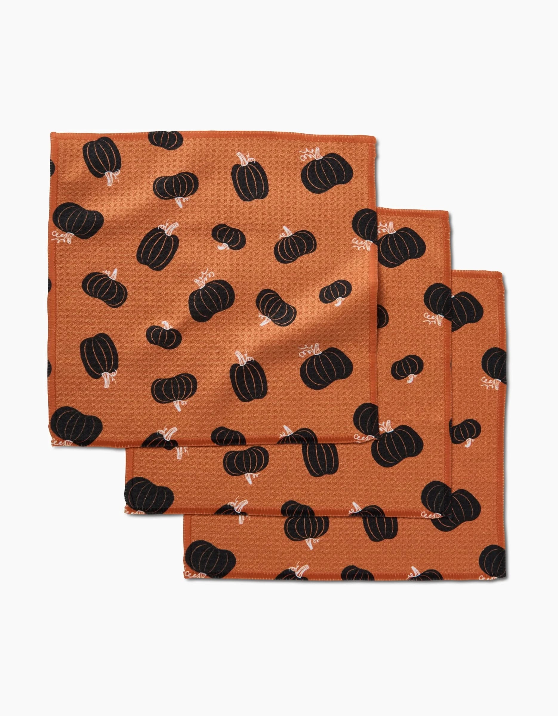 Three Halloween Black Pumpkins orange cloth napkins by Hufton Studio, made from recycled materials, display a repeating pattern of black pumpkins with white stems and are shown stacked and overlapping on a white background.
