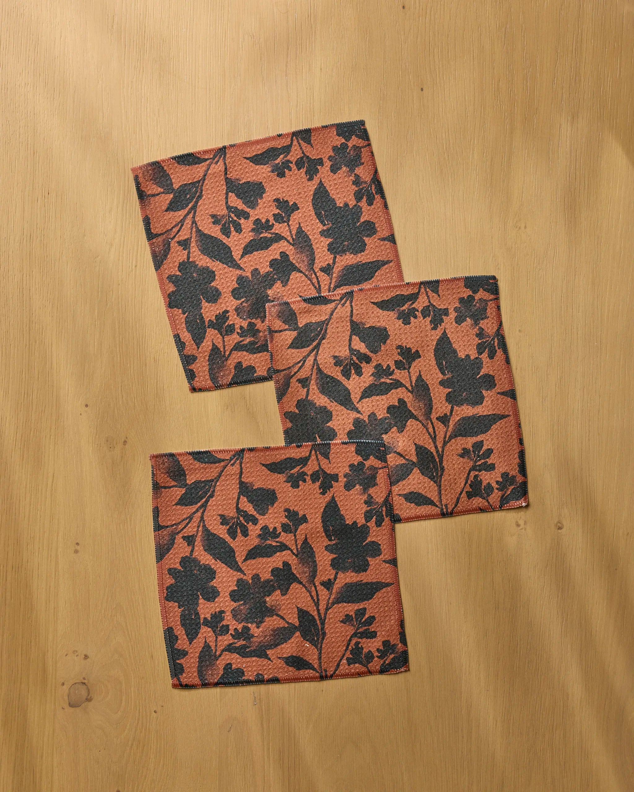 Three Hallowell square fabric napkins by Geometry, made from recycled materials and featuring a black floral print on reddish-brown, are arranged on a light wood surface.