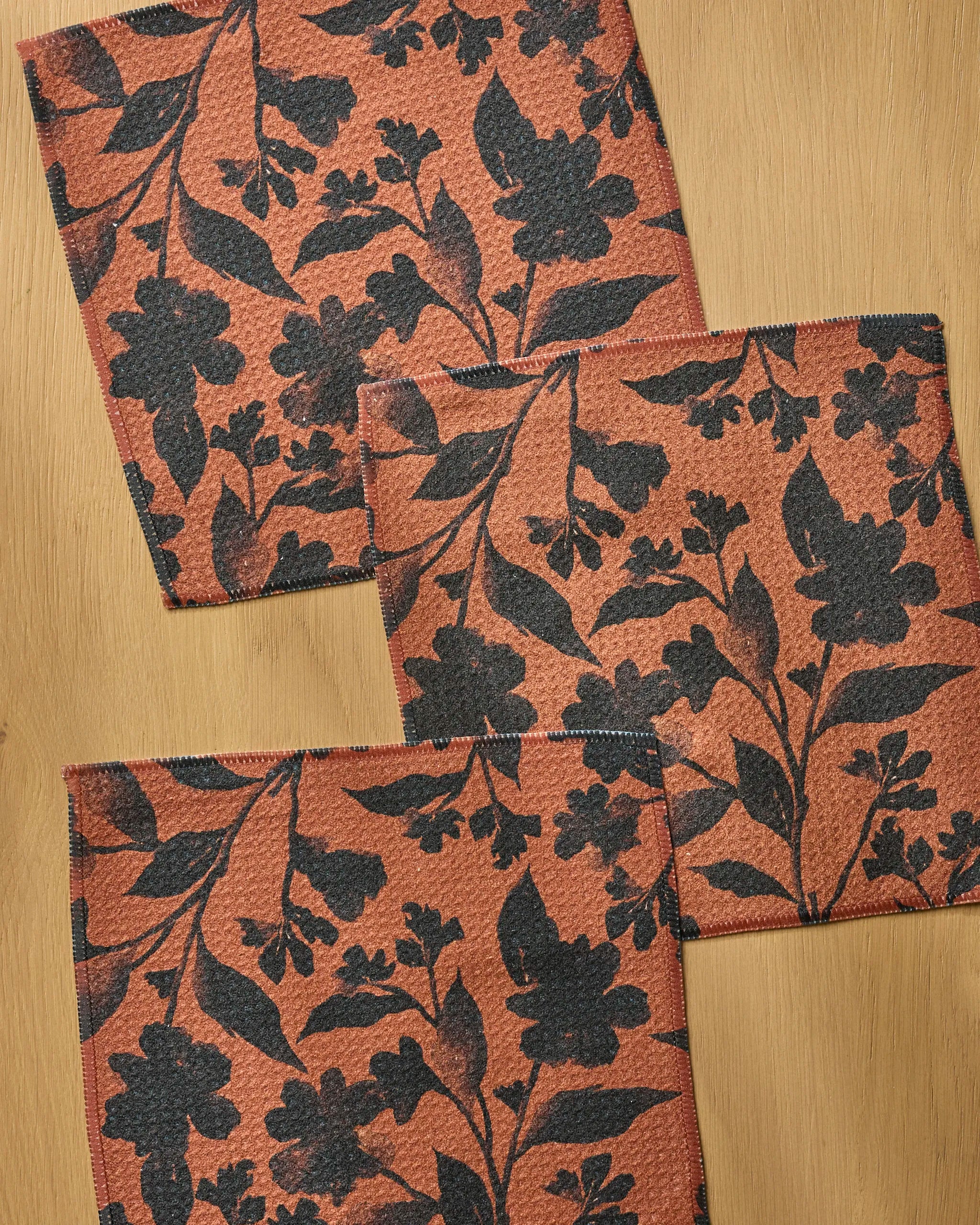 Four Hallowell napkins by Geometry, made from recycled materials and featuring a red and black floral design, are slightly overlapping on a light wooden surface.