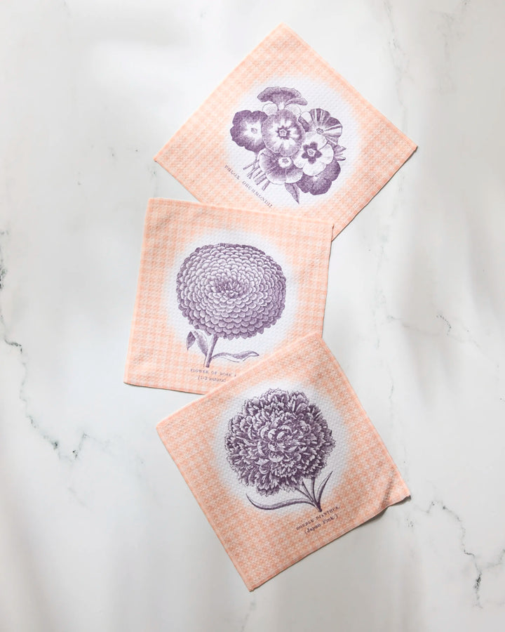 Three Geometry Hana peach dishtowels, featuring purple botanical flower designs and text, are arranged diagonally on a white marble surface.