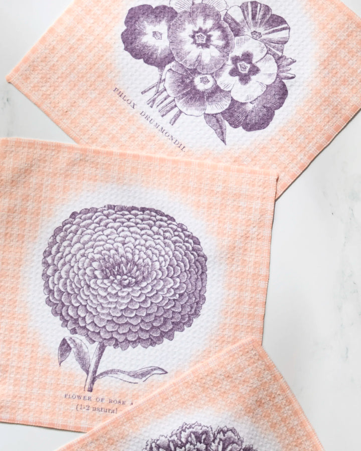 Three peach-colored Geometry Hana dishtowels with vintage-style purple flower designs—bouquet, chrysanthemum, and another bloom—are arranged diagonally on a white marble surface.