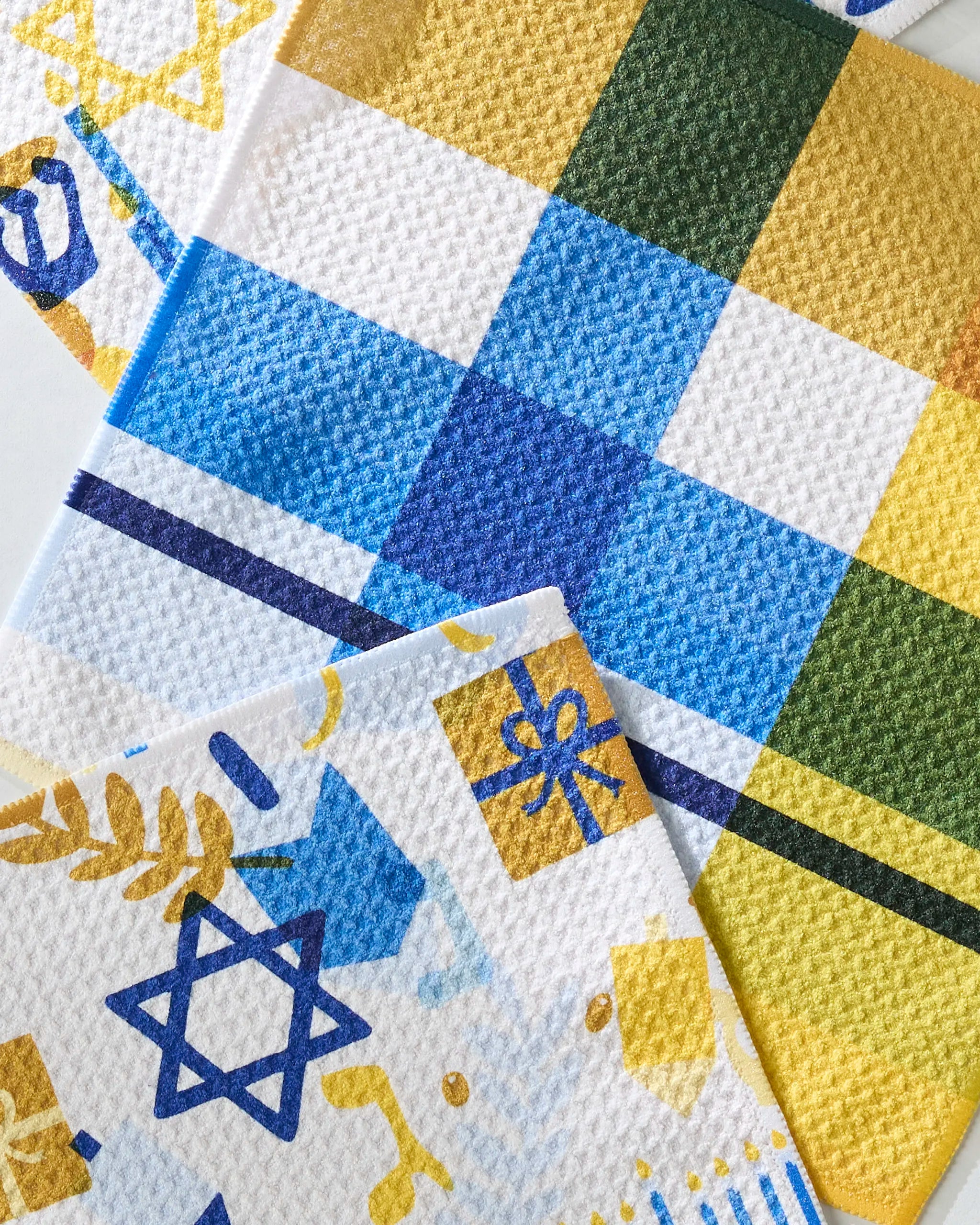 Close-up of Geometry’s “Hanukkah is Here” absorbent dishtowels, featuring festive blue and gold gift boxes, a Star of David, yellow menorahs, and a blue and yellow checkered background.