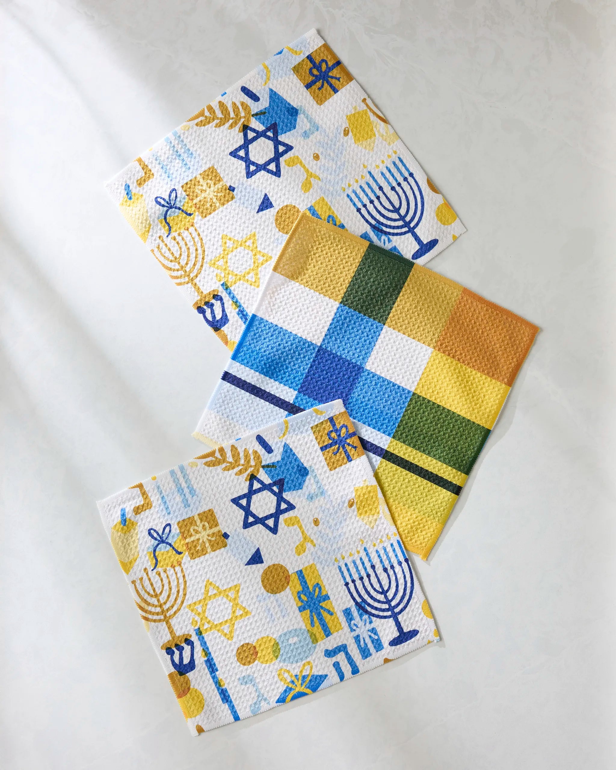 Three absorbent Geometry "Hanukkah is Here" dishtowels are displayed: two with menorahs, Stars of David, and dreidels in blue and gold, and one with a simple blue, yellow, and green checkered pattern.