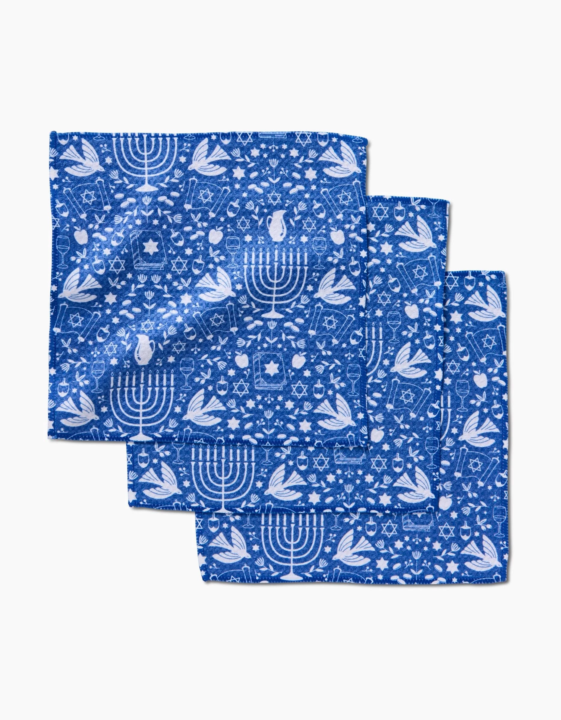 Three blue Geoweave cloth napkins from Megan Kampa’s Hanukkah Sameach collection, featuring white Hanukkah-themed designs—menorahs, dreidels, doves, and Stars of David—are neatly folded and stacked on a white background.