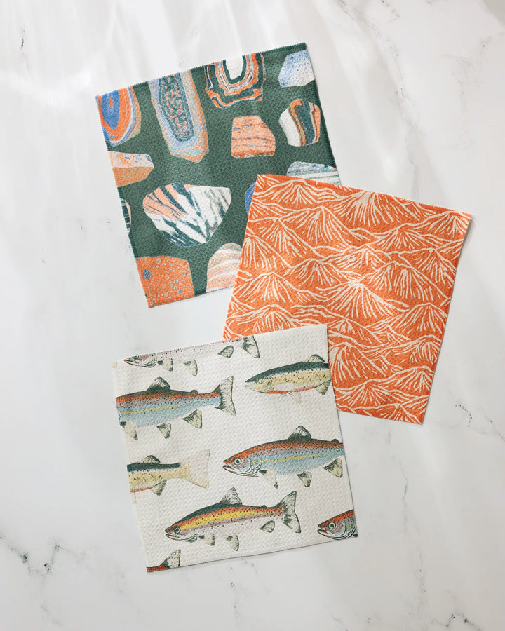 Three Geometry Happy Camper reusable dishcloths rest on a white marble surface: one with colorful abstract shapes, one orange with a leaf pattern, and one covered in fish illustrations.