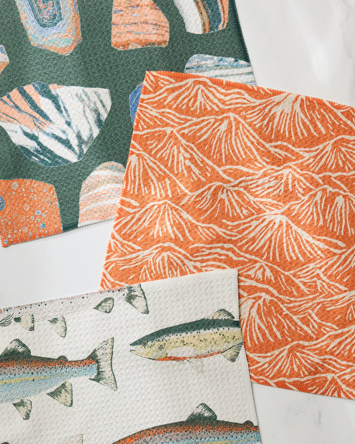 The Geometry Happy Camper set features three vibrant reusable dishcloths: orange and green stones and shells, orange and white mountain lines, and green fish on white—all displayed on a white surface.