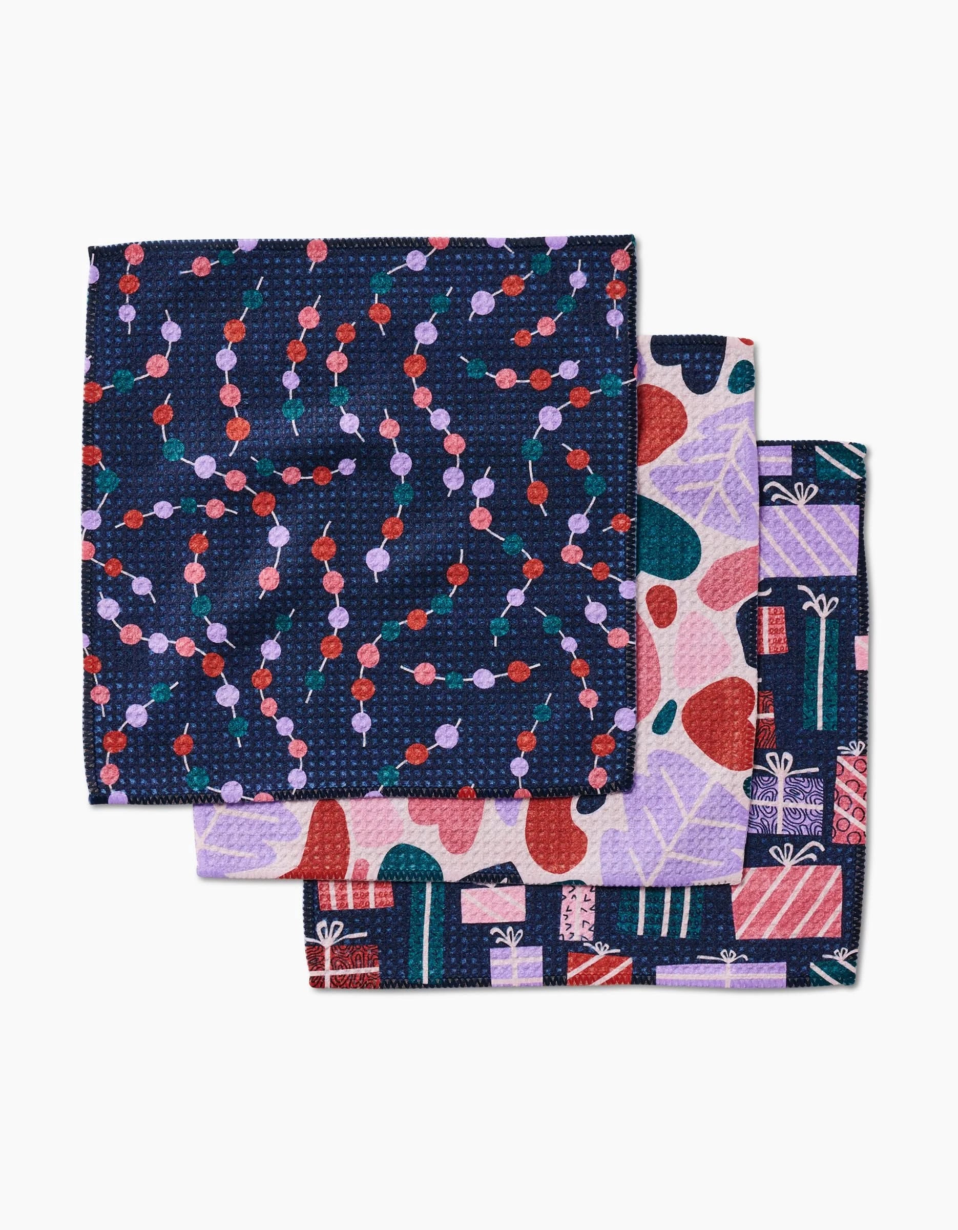 Three folded Happy Holly cloth napkins by Claire Iglesias: the top shows a navy base with red and purple dots, the second has multicolored shapes, and the third features gift boxes and abstract patterns. Made from Geoweave fabric.