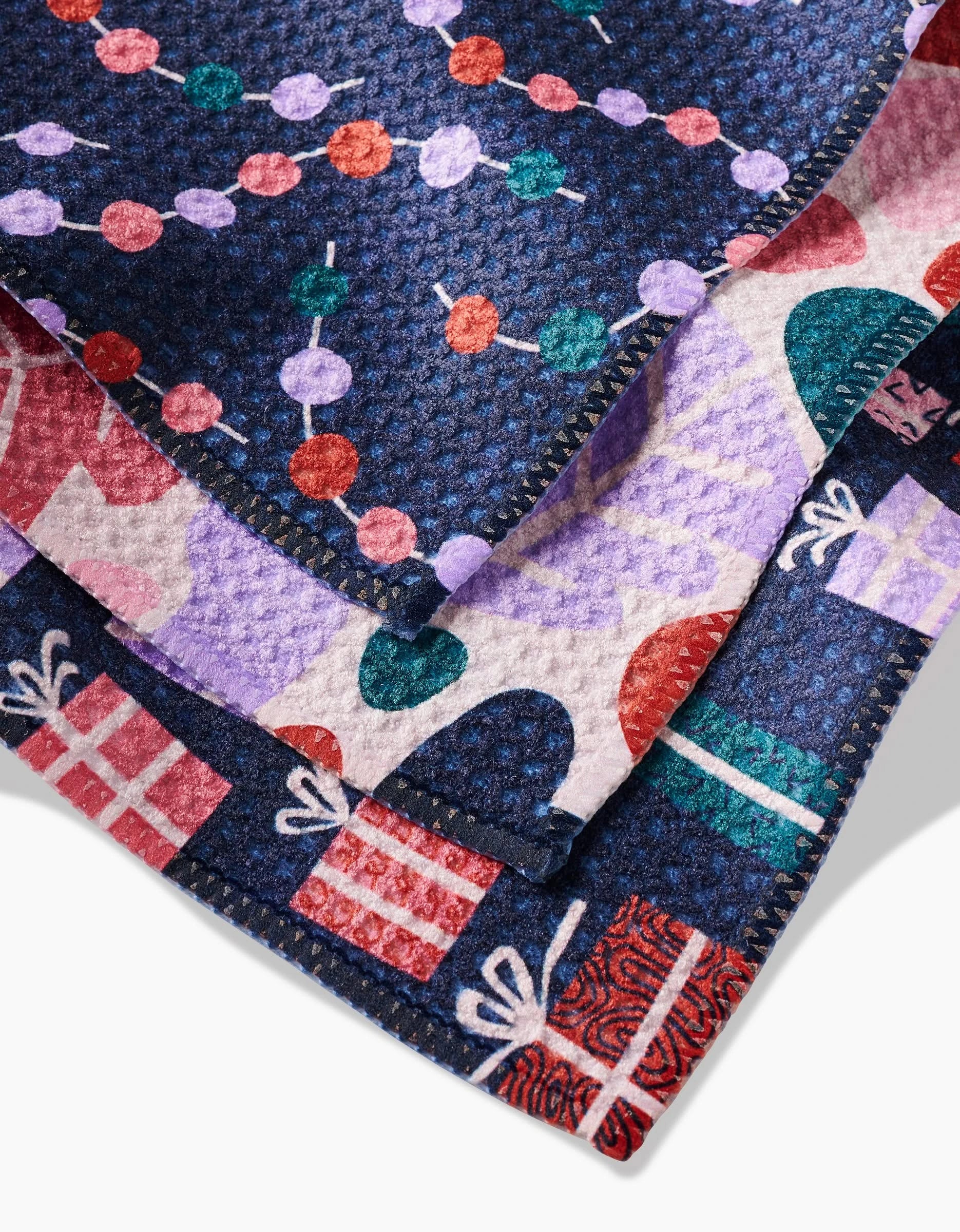 A close-up of the Claire Iglesias Happy Holly dishcloth shows its colorful Geoweave fabric with festive patterns of wrapped gifts, ornaments, and holiday lights in red, purple, green, and blue.