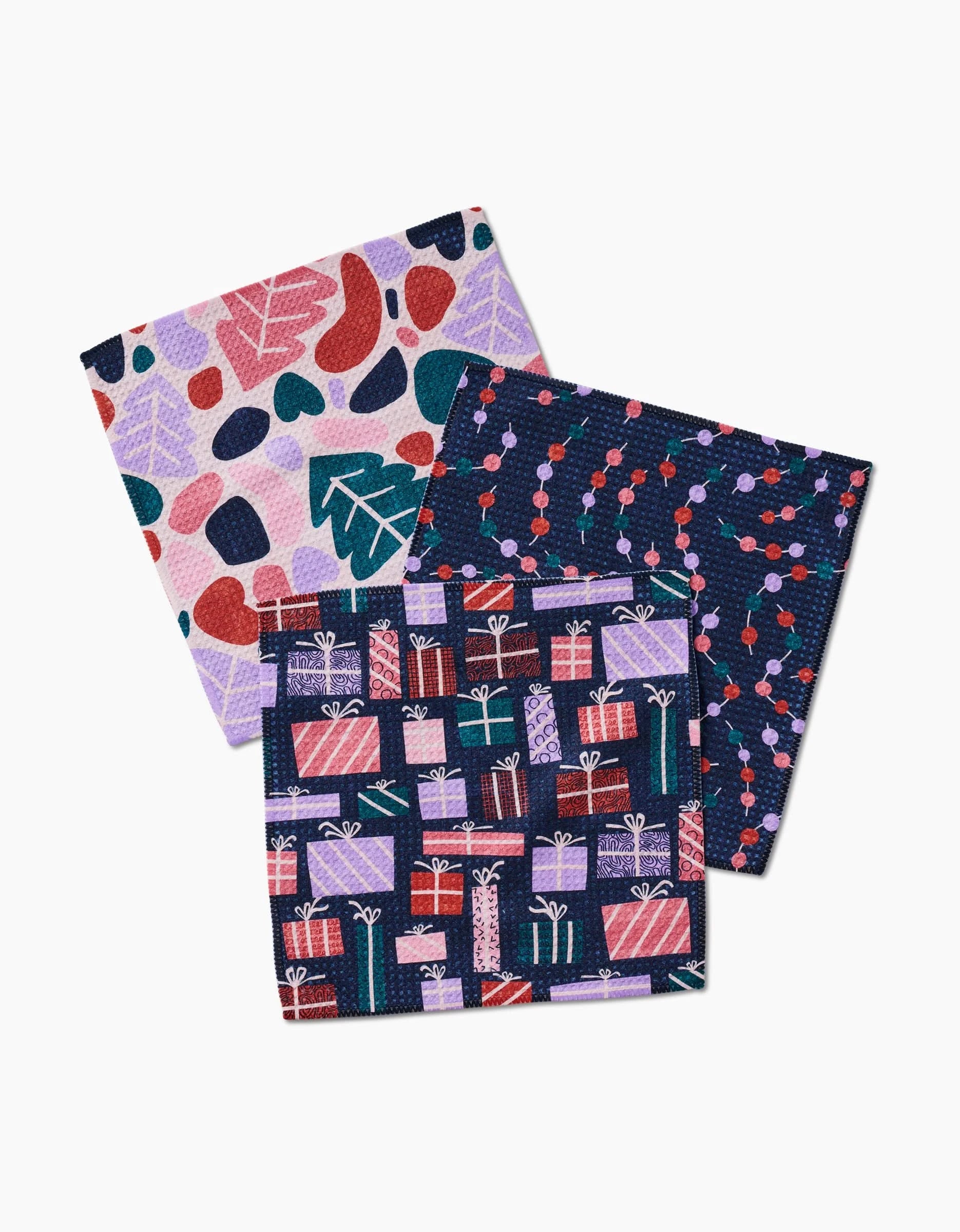 The Claire Iglesias Happy Holly set includes three colorful Geoweave dishcloths: one with abstract leaf and bean shapes, one with red and purple berries on dark blue, and one featuring festive gift box illustrations.