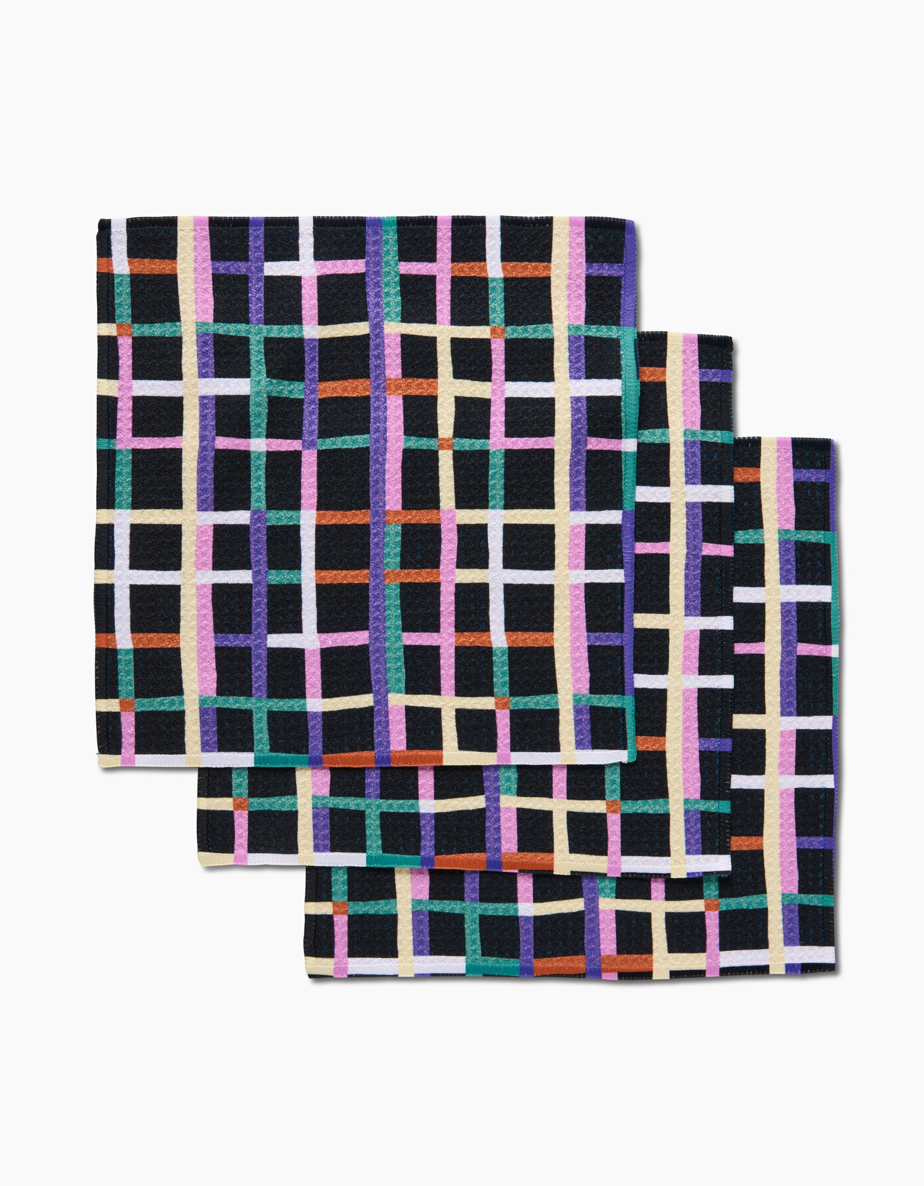 Three folded Happy Plaid napkins by Claire Iglesias, made from Geoweave fabric, display a colorful grid of purple, orange, green, blue, pink, and cream lines on black, arranged diagonally on a white surface.