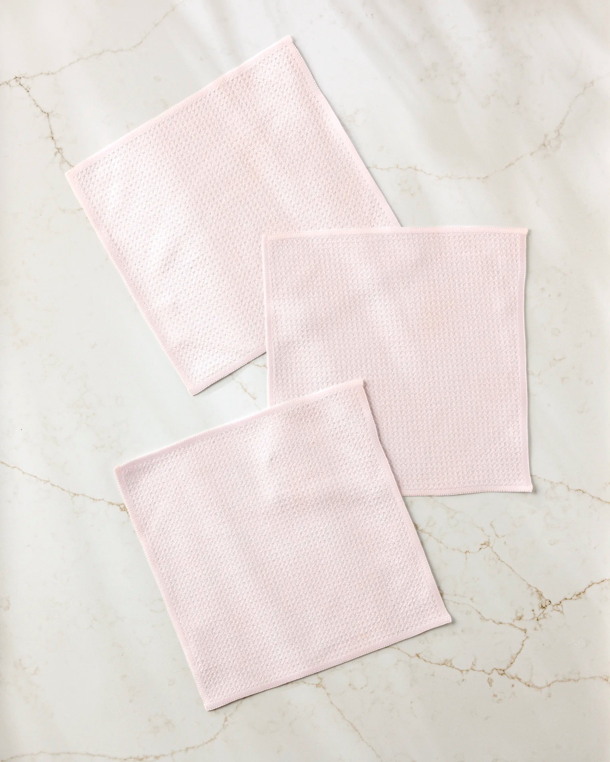 Three Harper Pink textured cloth squares from Kailee Wright's super-absorbent dishcloth set are neatly arranged on a white marble surface with light gray and beige veining.