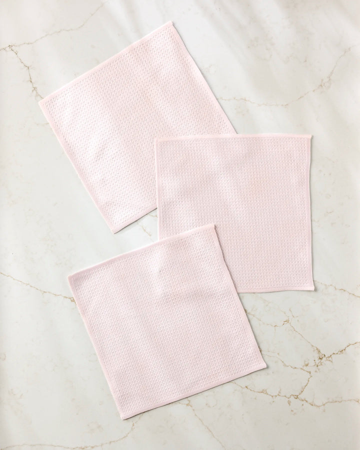 Three Harper Pink textured cloth squares from Kailee Wright's super-absorbent dishcloth set are neatly arranged on a white marble surface with light gray and beige veining.