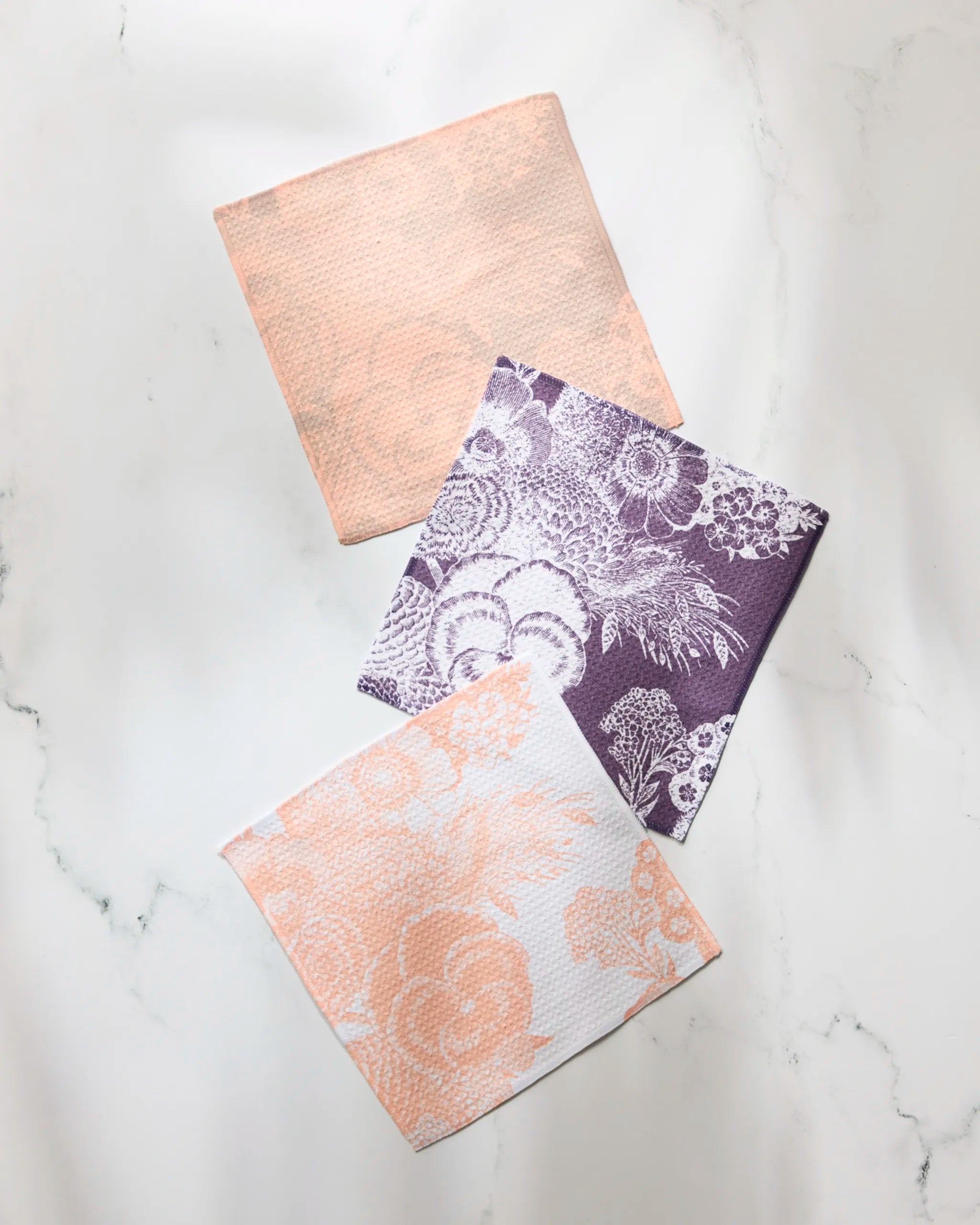 Three textured cloths from Geometry’s Haru fast-drying dishcloth set are displayed on white marble: one solid peach, one purple with white floral lace, and one white featuring peach floral designs.