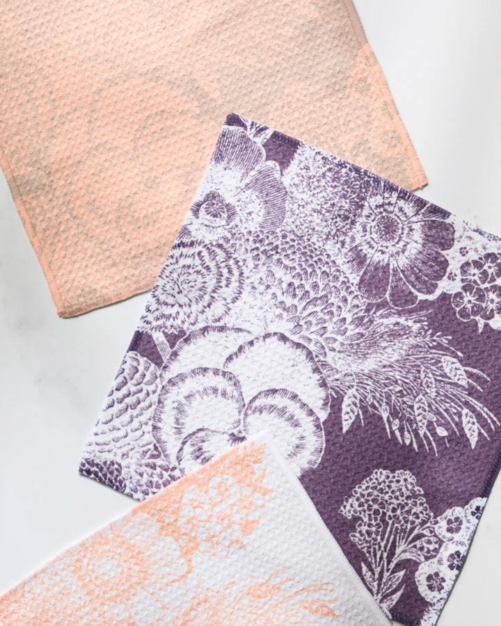The Geometry Haru set features three textured recycled cloths: one solid peach, one white with peach floral patterns, and one purple with white floral and botanical illustrations, all arranged on a white surface.