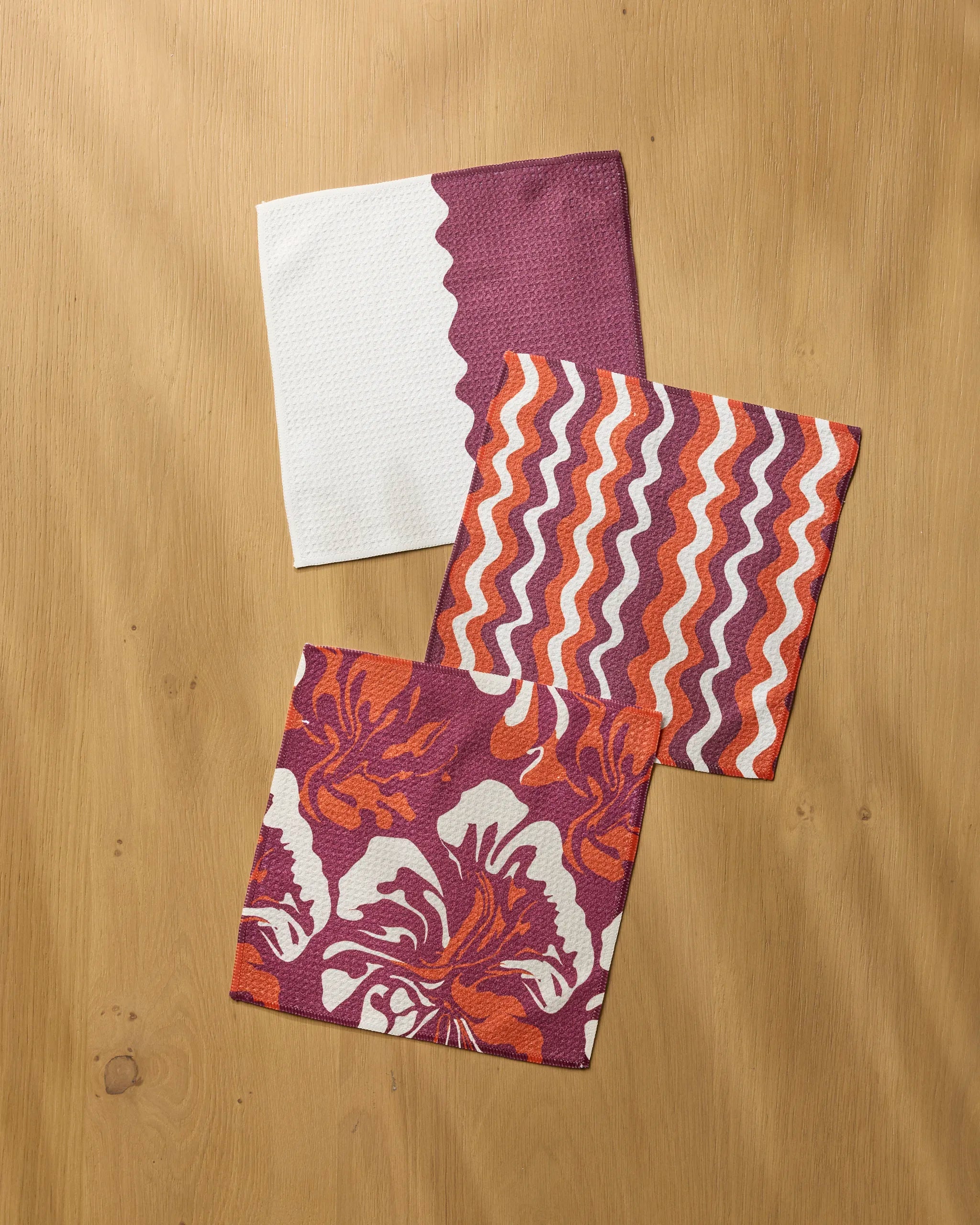 Three colorful napkins from Geometry’s Harvest Groove Muse set rest on a wooden surface: one is white with a purple zigzag edge, one sports wavy orange and purple stripes, and one displays a purple, orange, and white floral pattern.