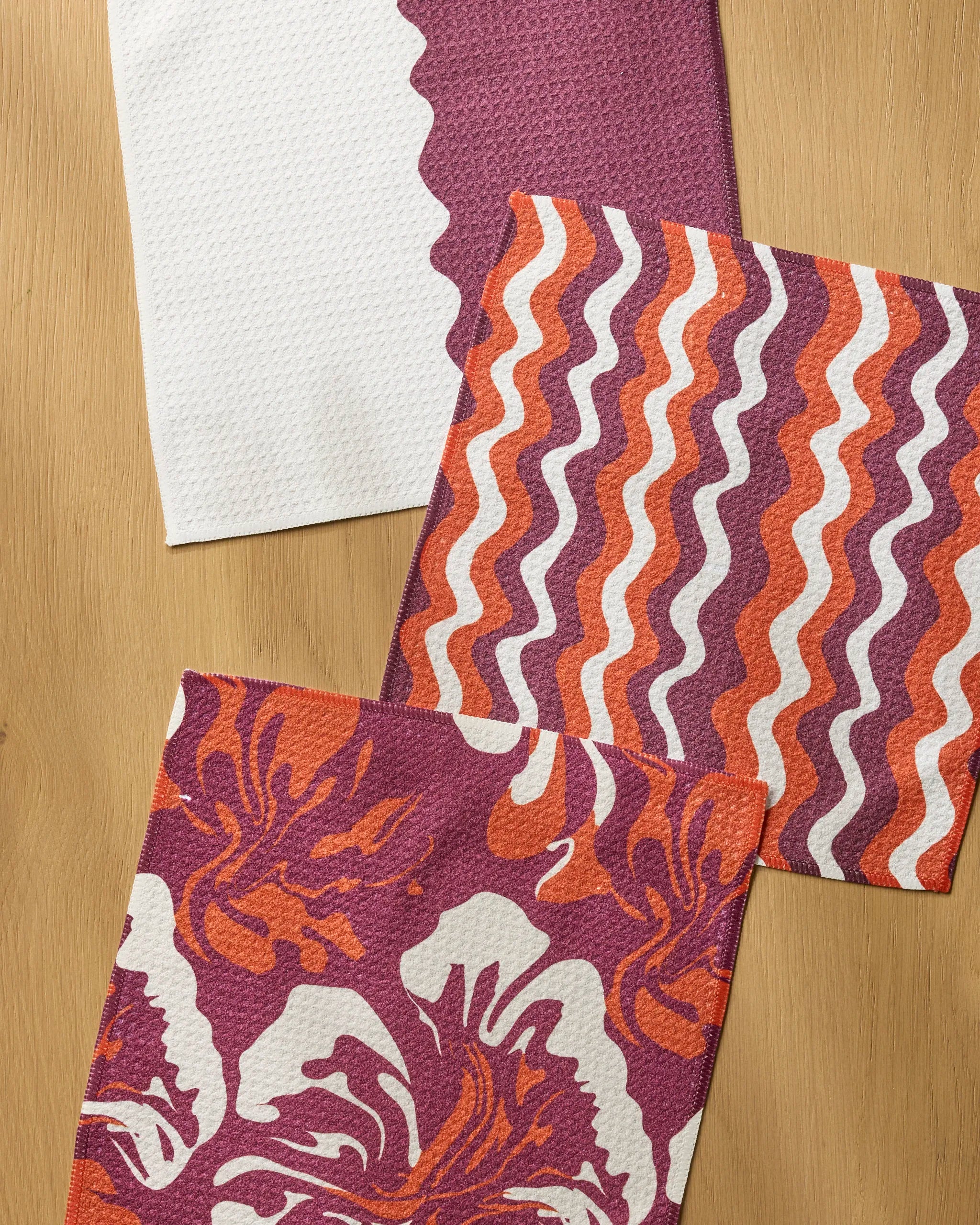 The Geometry Harvest Groove Muse set features three textured cloth napkins—a white with purple scalloped edge, a striped orange-purple-white, and a bold floral print—ideal as absorbent dishcloths for any kitchen.