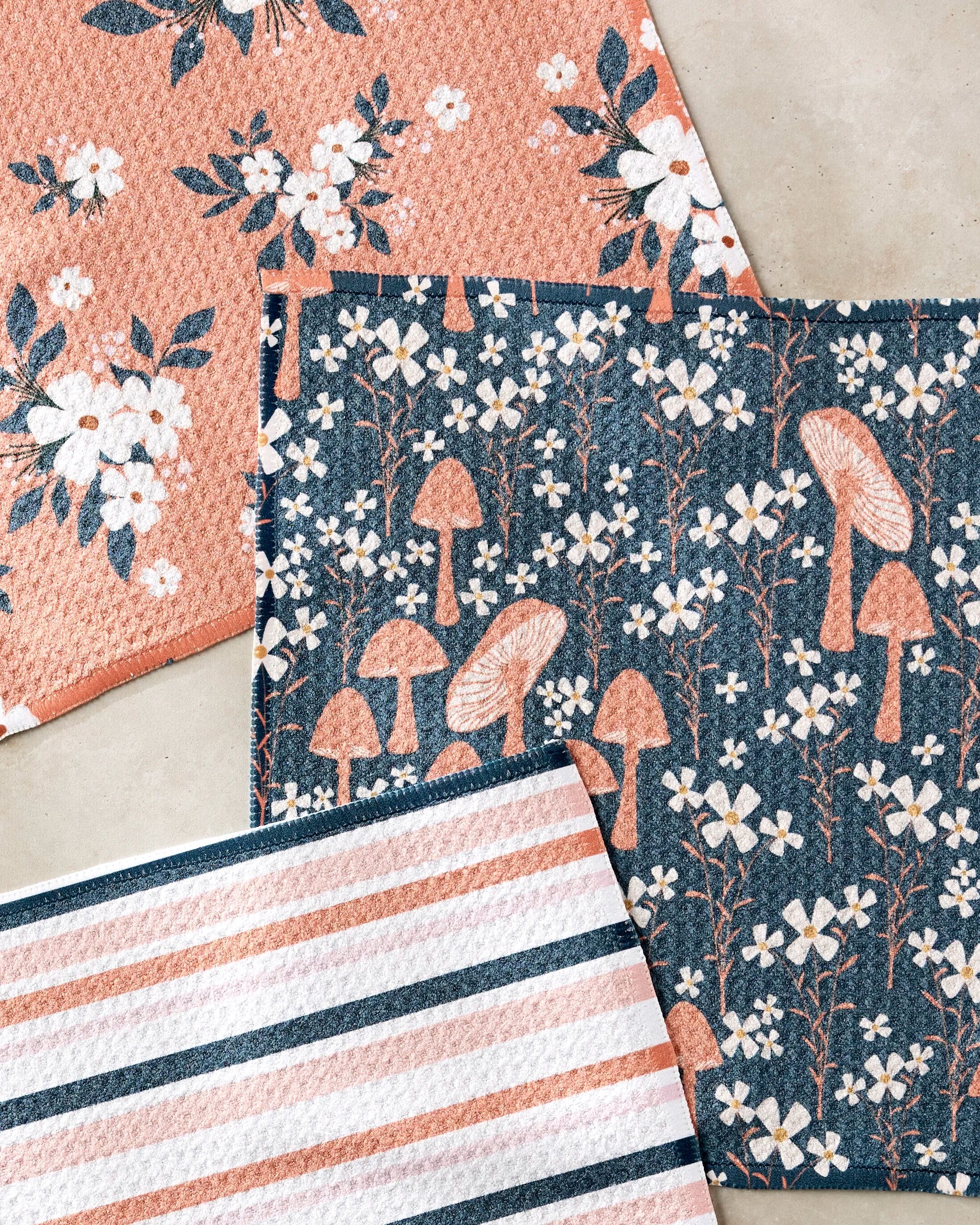 The Harvest Season dishcloth set by Charlie Rowan Designs includes three reusable cloths: white flowers on peach, mushrooms and white flowers on blue, and peach, blue, and white stripes.