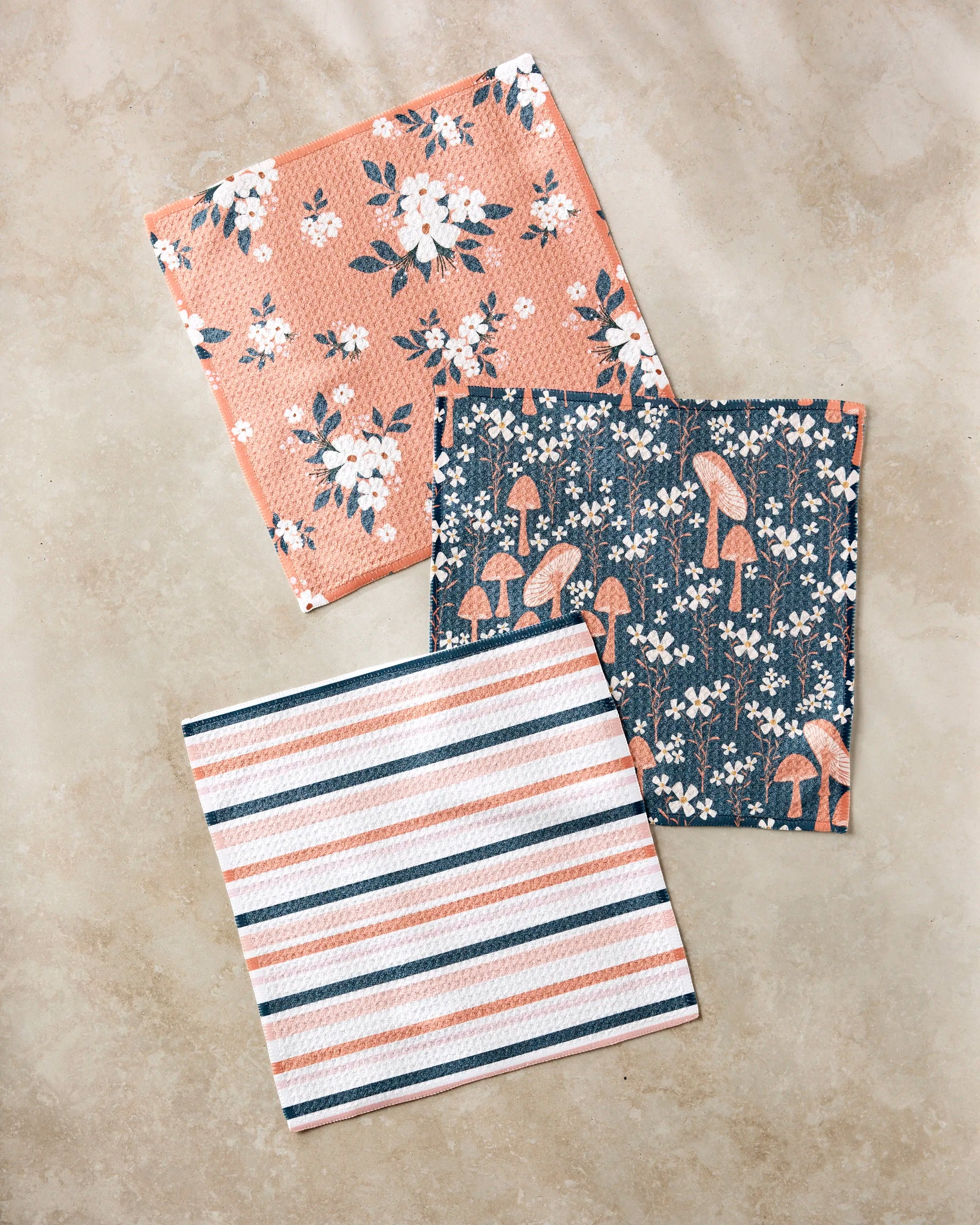 The Harvest Season dishcloth set by Charlie Rowan Designs features three reusable cloths: pink floral, blue with mushrooms and birds, and striped patterns—an eco-friendly kitchen essential.