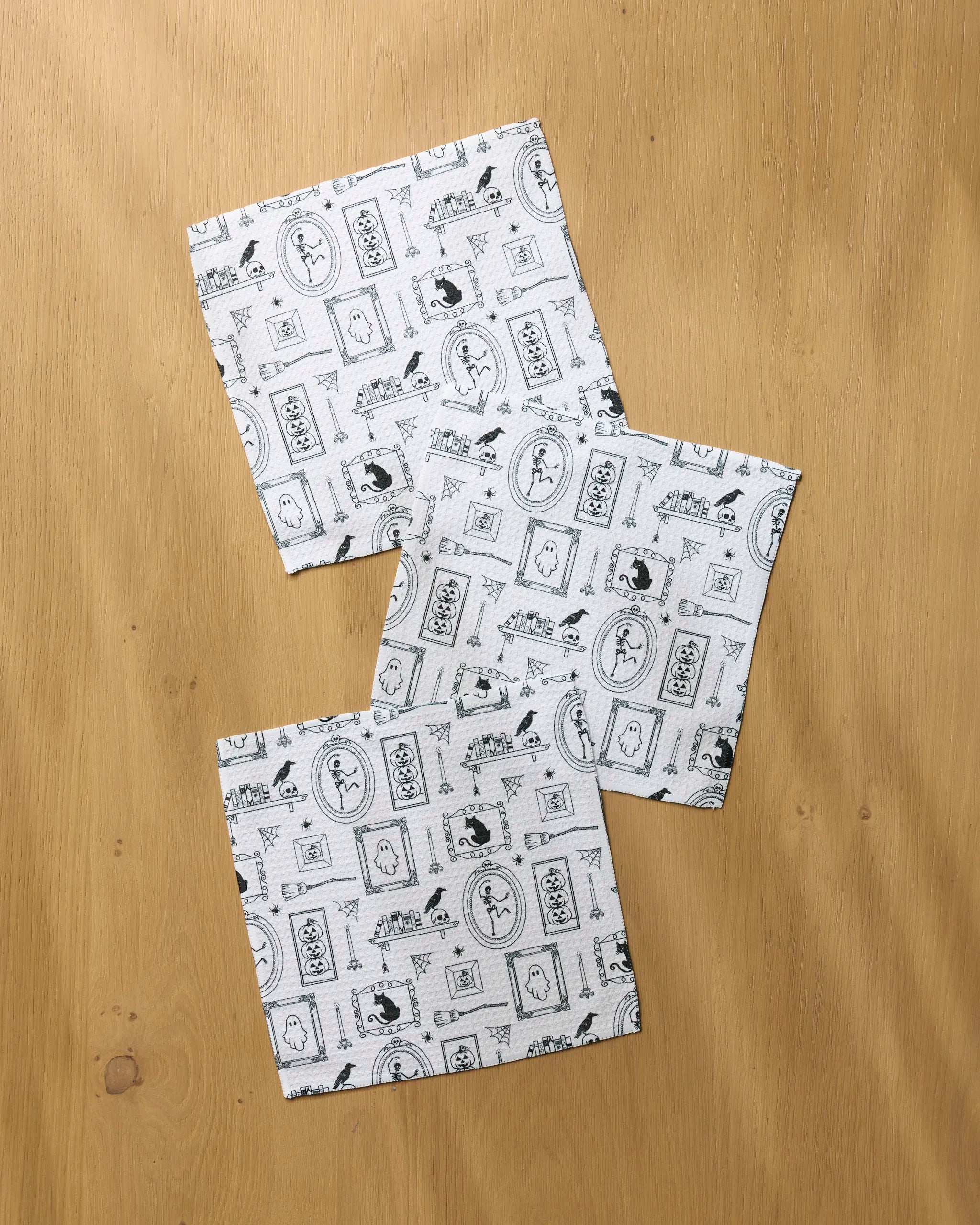 Three square white "Haunted Gallery" dishcloths by Geometry, featuring black printed motifs such as circles, rectangles, and animal shapes, overlap slightly and are casually spread on a light wood surface.