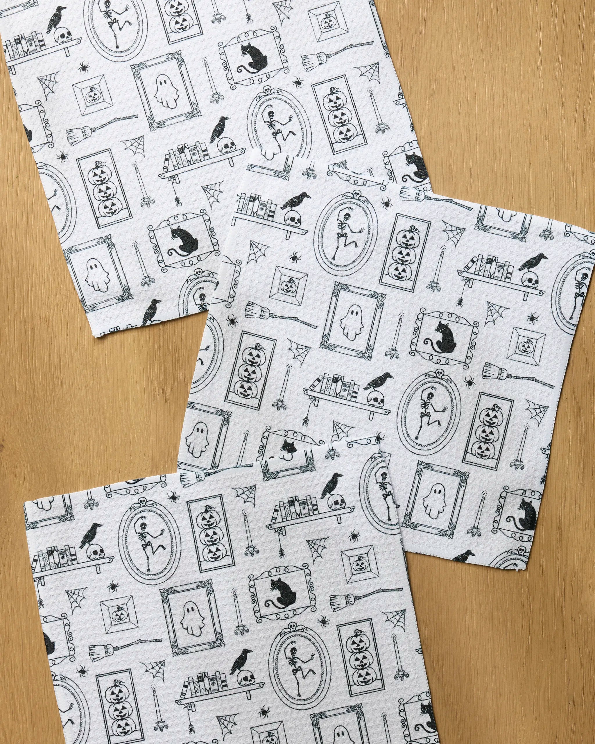 The Geometry Haunted Gallery set features three white recycled dishcloths with black Halloween-themed prints—ghosts, pumpkins, spiders, bats, and portraits—displayed on a wooden surface for a festive touch.
