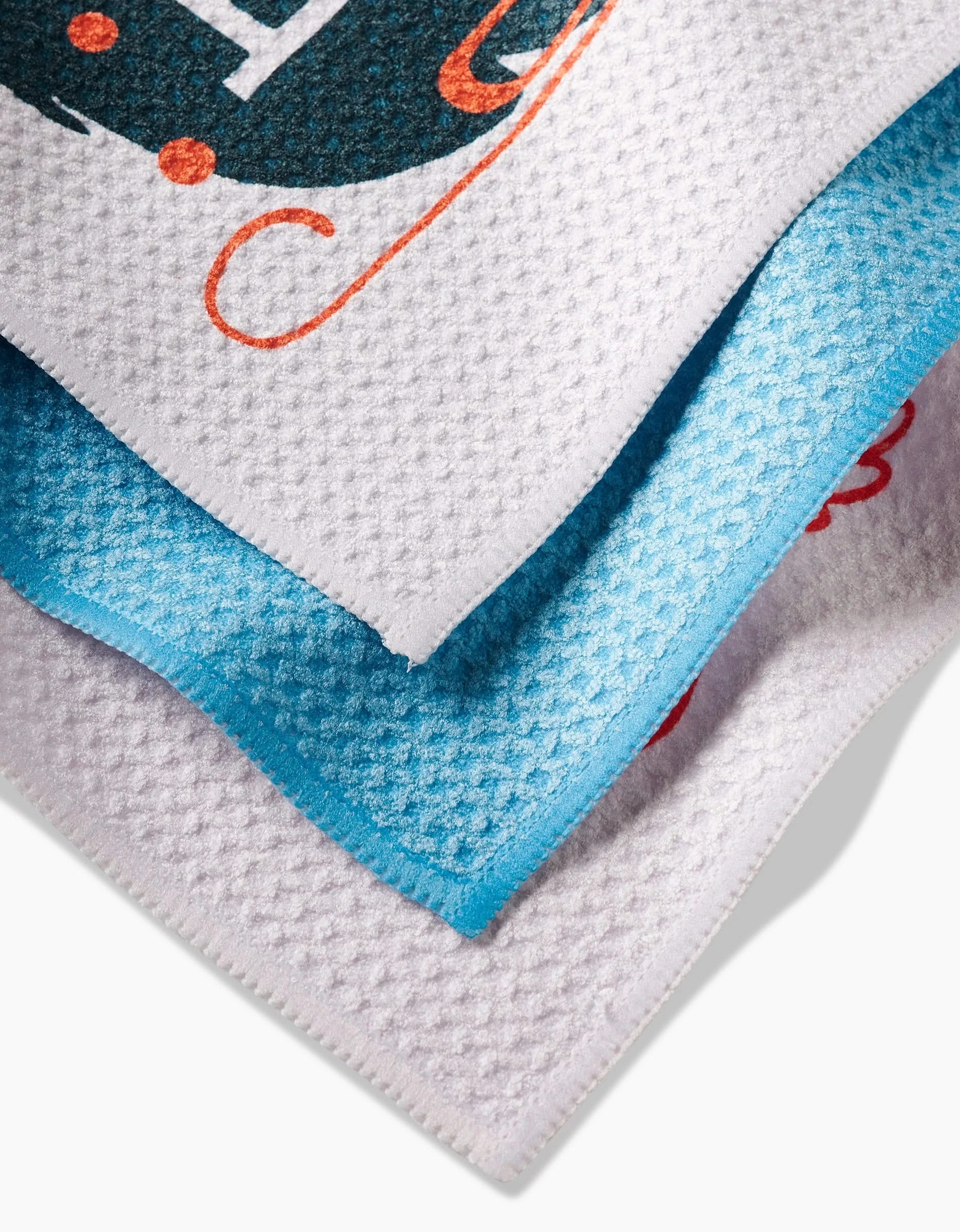 Three Carly Berry Here Comes Santa Claus microfiber towels in white and blue, stacked diagonally, showcase Geoweave fabric with partial colorful festive prints visible on the top and bottom towels.