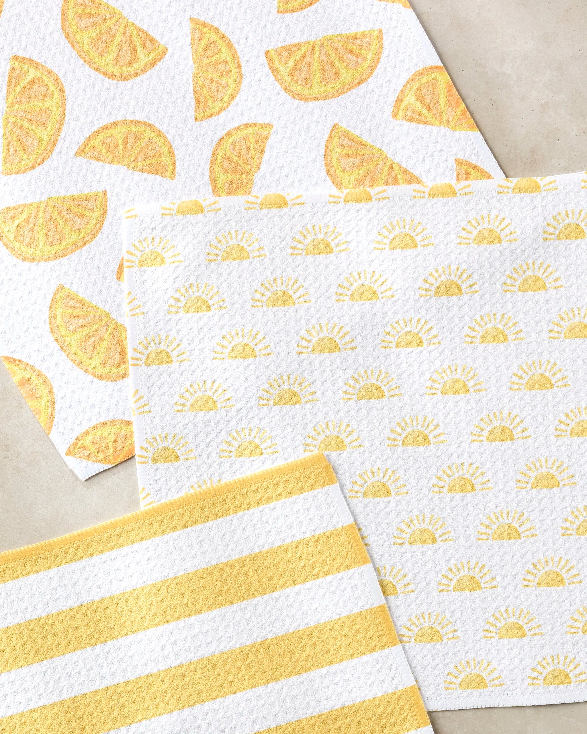 Three Aubrey Fairchild "Here Comes The Sun" dishtowels are shown: one with lemon wedges, one with sun shapes, and one with yellow and white stripes, all arranged on a light surface.
