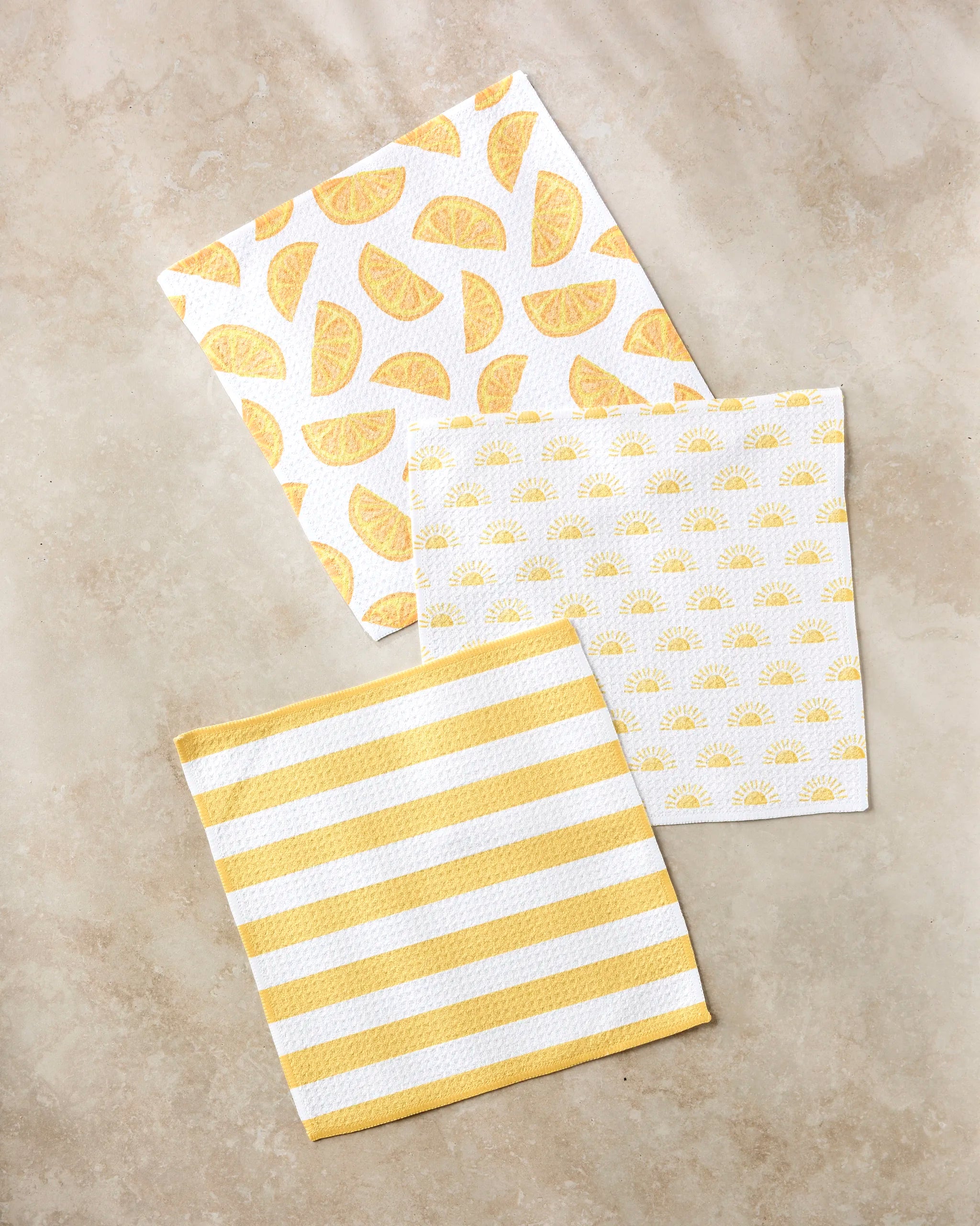 Three "Here Comes The Sun" kitchen towels by Aubrey Fairchild are arranged on a beige surface: one with lemon wedges, one with small suns, and one featuring yellow and white stripes.