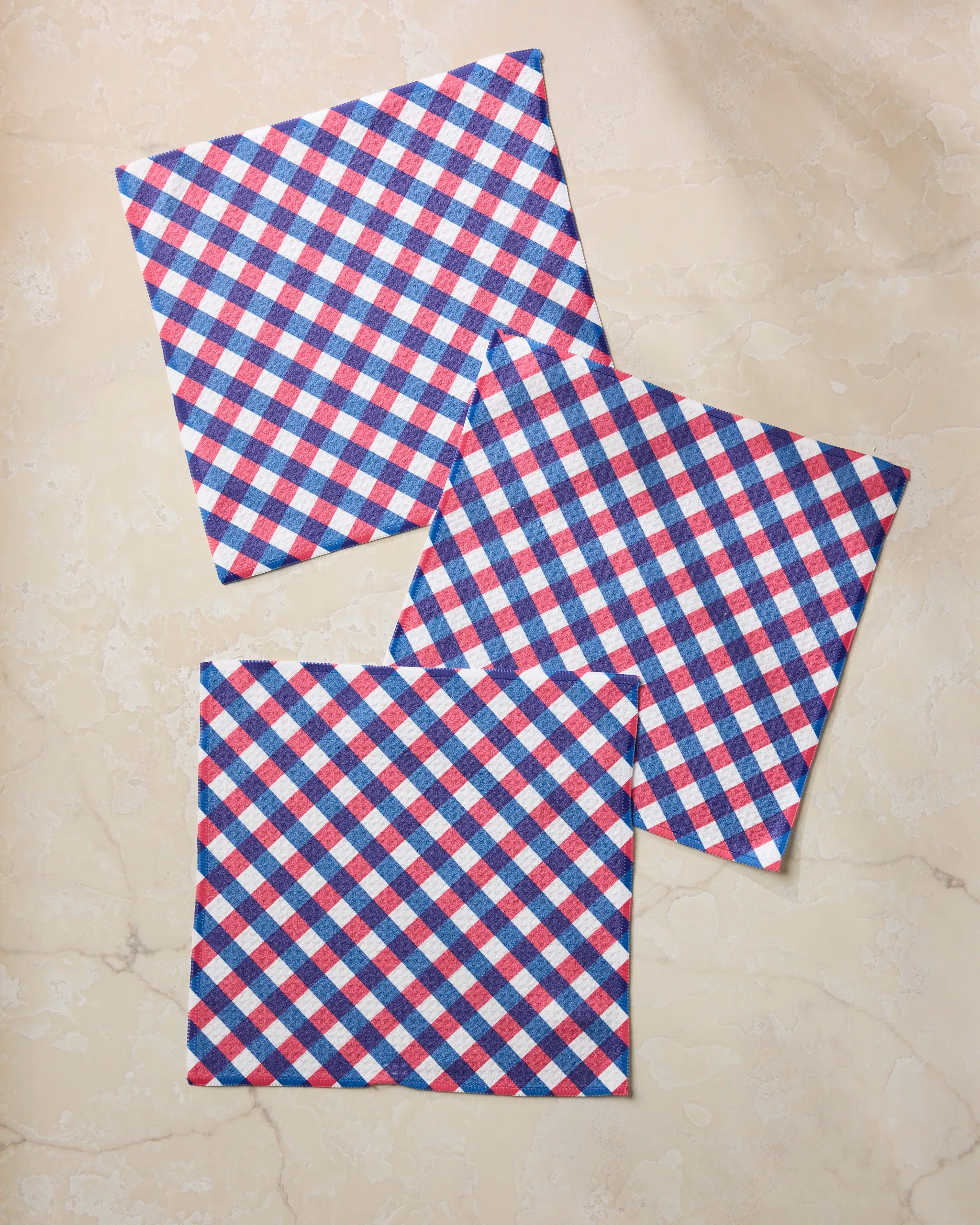 Three Heritage Check napkins by Geometry, featuring a red, white, and blue checkered pattern and made from recycled kitchen towels, are arranged with slight overlap on a light marble surface.