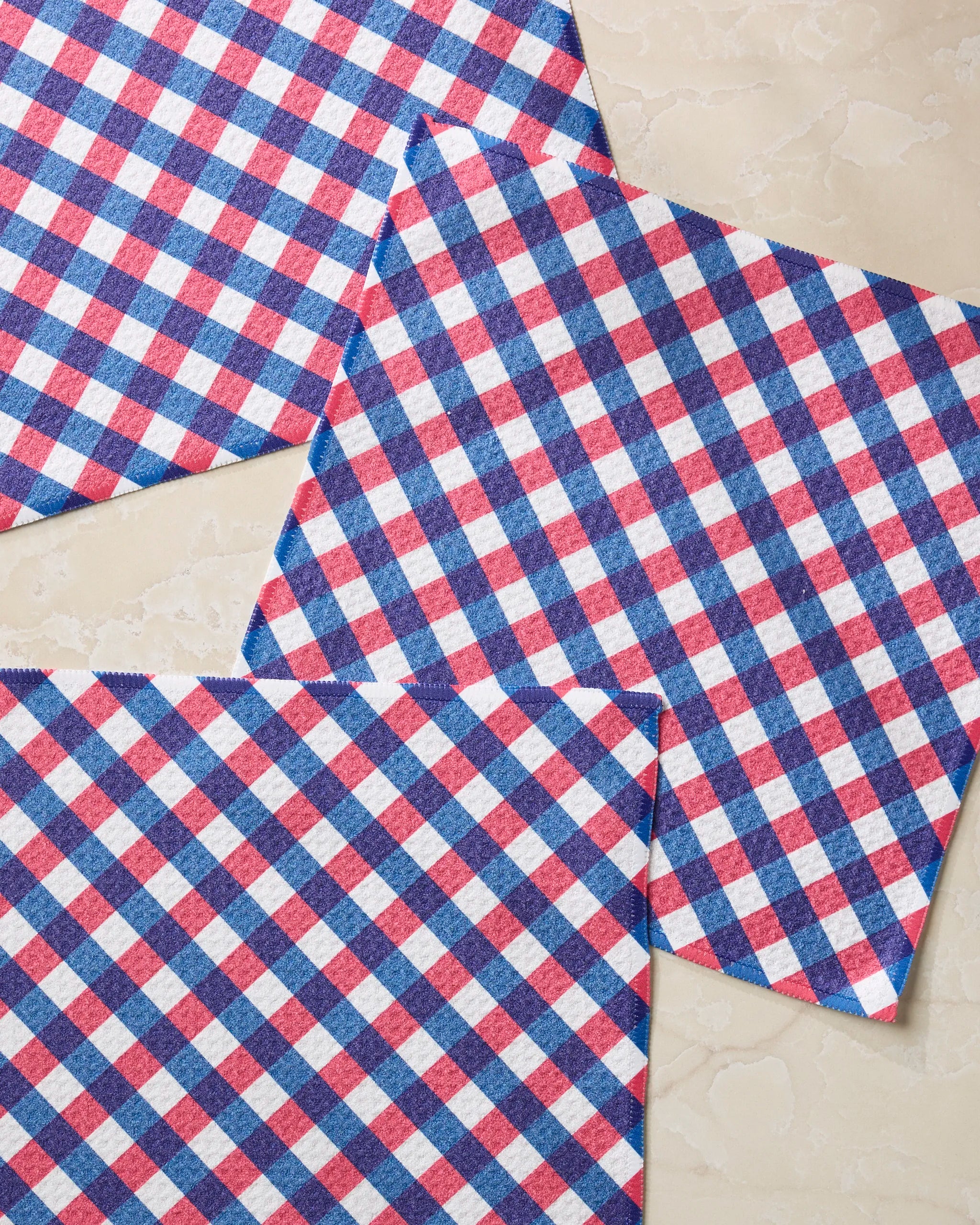 Three Geometry Heritage Check dish towels, featuring a red, white, and blue check pattern, are arranged overlapping at varied angles on a light marble surface.