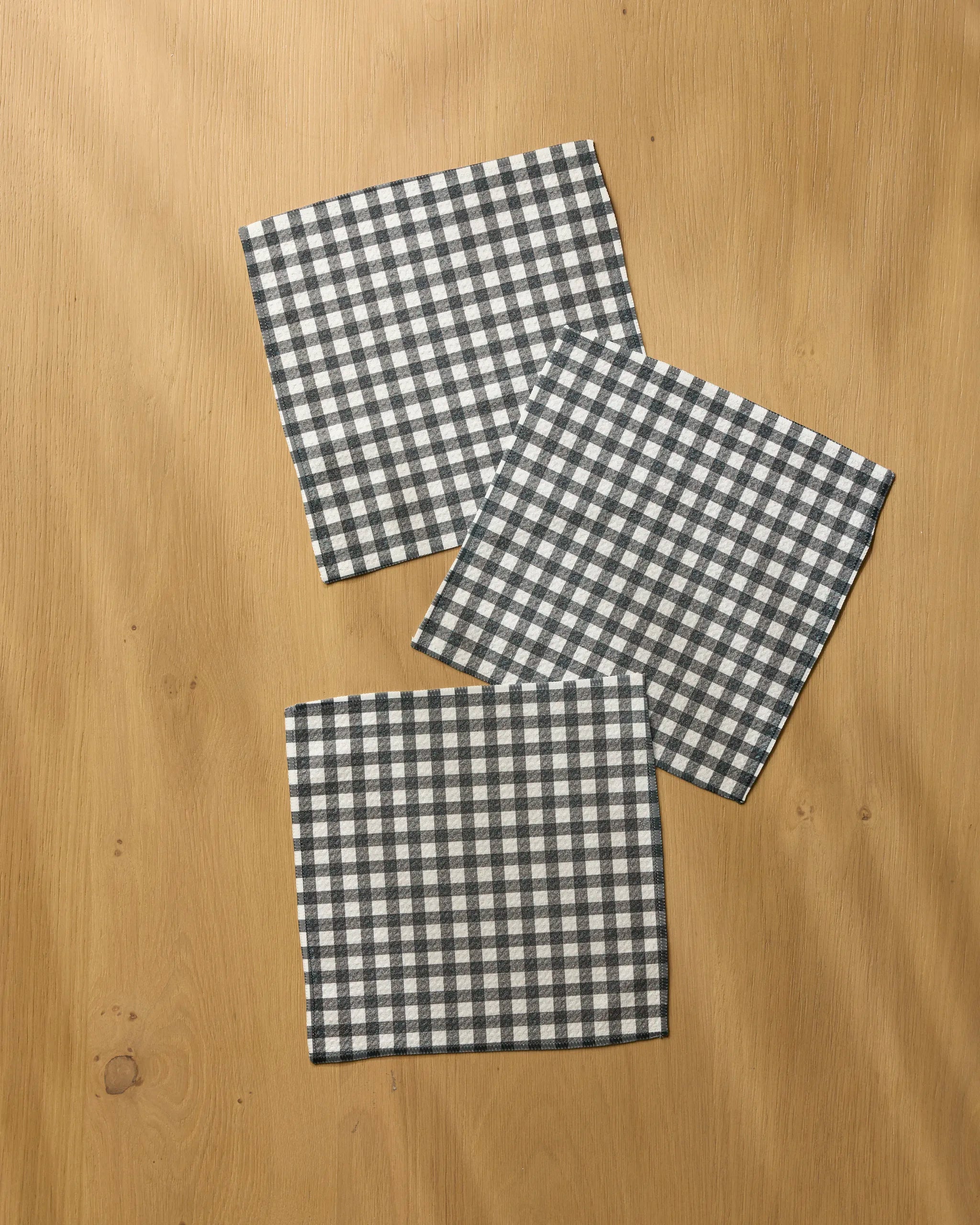 Three Heritage cloth napkins from Geometry, featuring a black and white checkered pattern and super-absorbent design, are neatly arranged in a scattered layout on a light wooden surface.