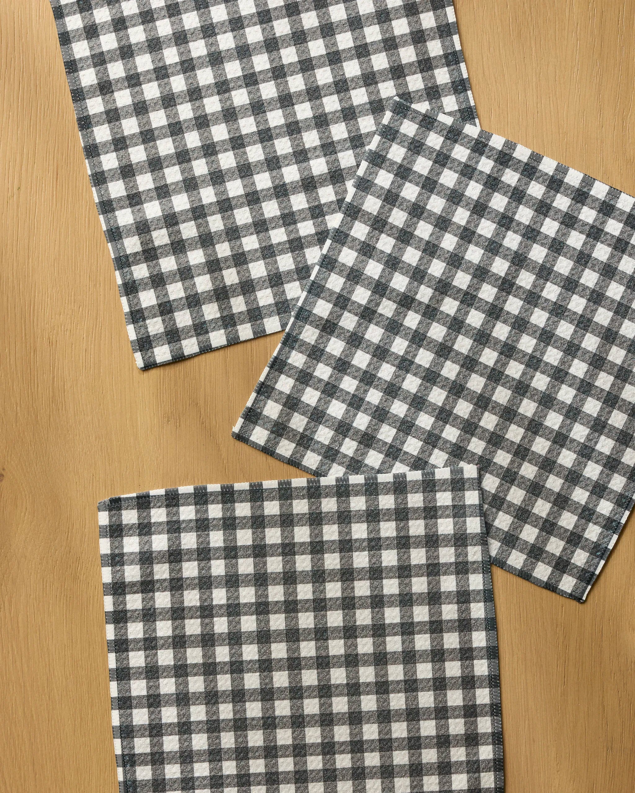 Three "Heritage" napkins by Geometry, featuring a black and white checkered design and made from recycled materials, are arranged at varying angles atop a light wooden surface.