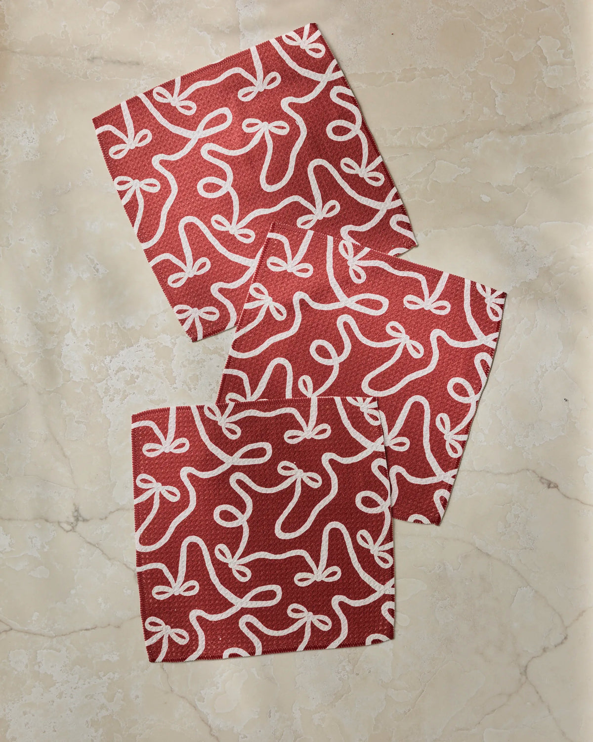 Three “Holiday Bows” napkins by Geometry, featuring a red design with white bow patterns and made from recycled materials, are arranged on a beige marble surface, each slightly overlapping the next.