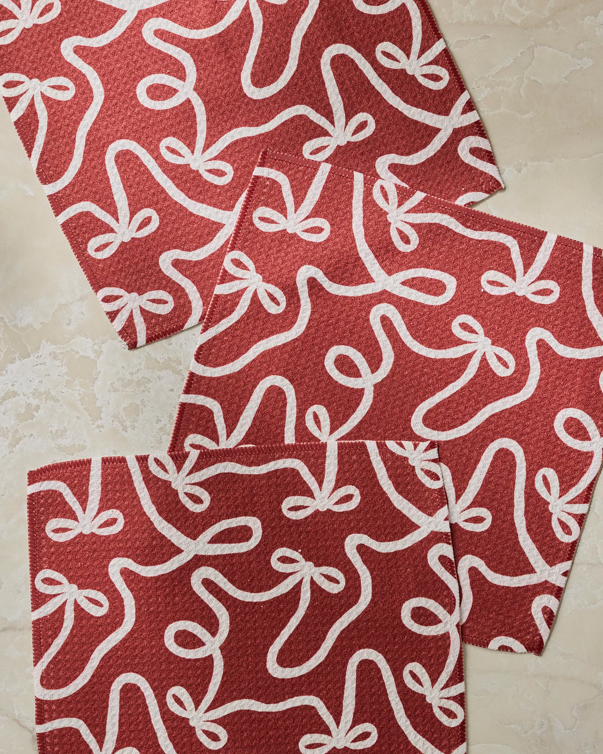 Three red rectangular “Holiday Bows” papers by Geometry, featuring white ribbon-like loop patterns, overlap on a beige marble surface.