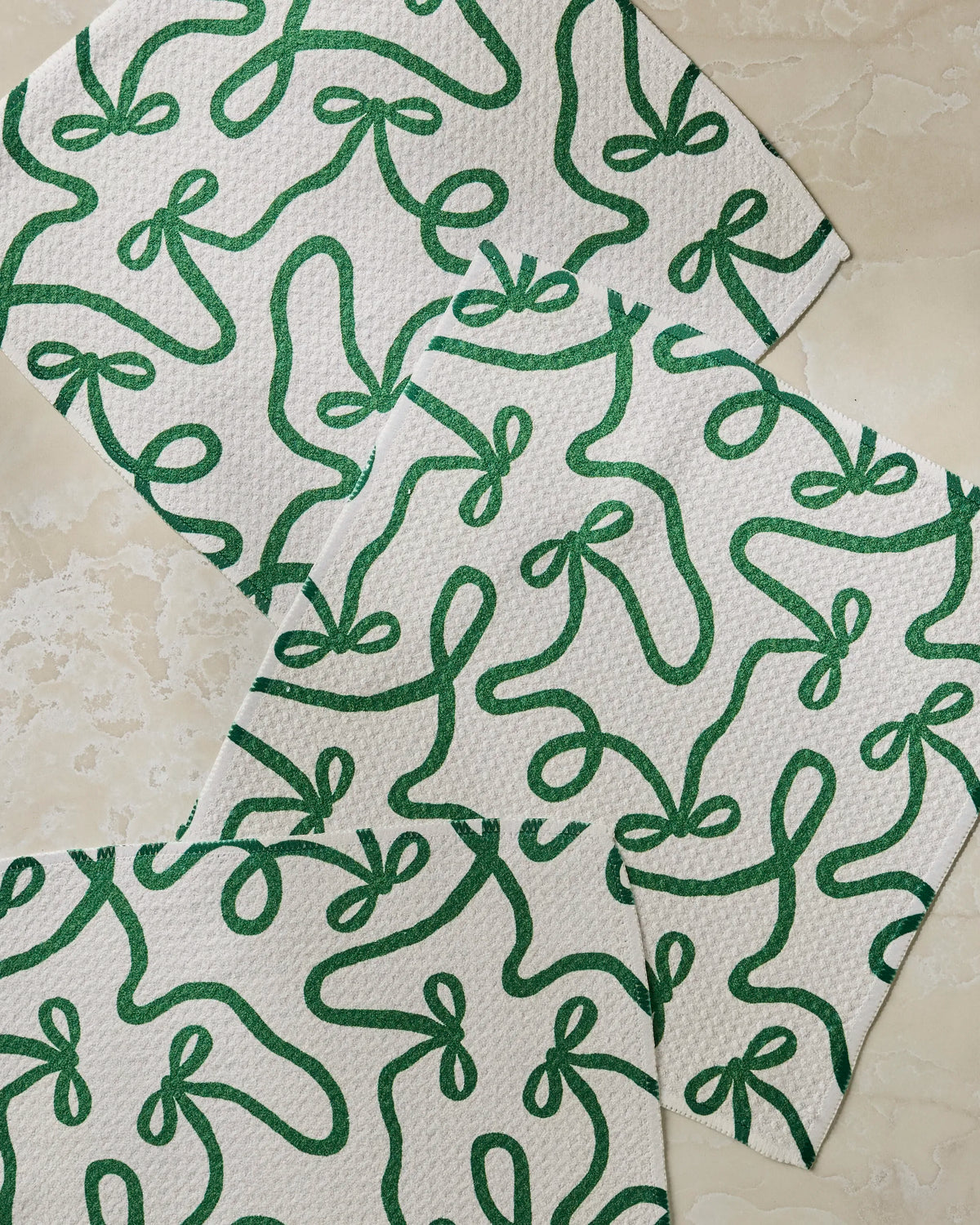 Four “Holiday Bows” napkins by Geometry, featuring a green abstract ribbon design on white textured fabric made from recycled materials, are arranged in a slight overlap on a light marble surface.