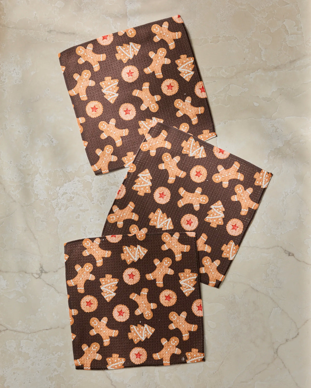Three “Holiday Cookies” cloth napkins by Geometry, crafted from recycled materials and adorned with gingerbread men, Christmas trees, and star cookie designs, are arranged on a light marble surface. This festive set blends eco-friendly practicality with cheerful holiday style.