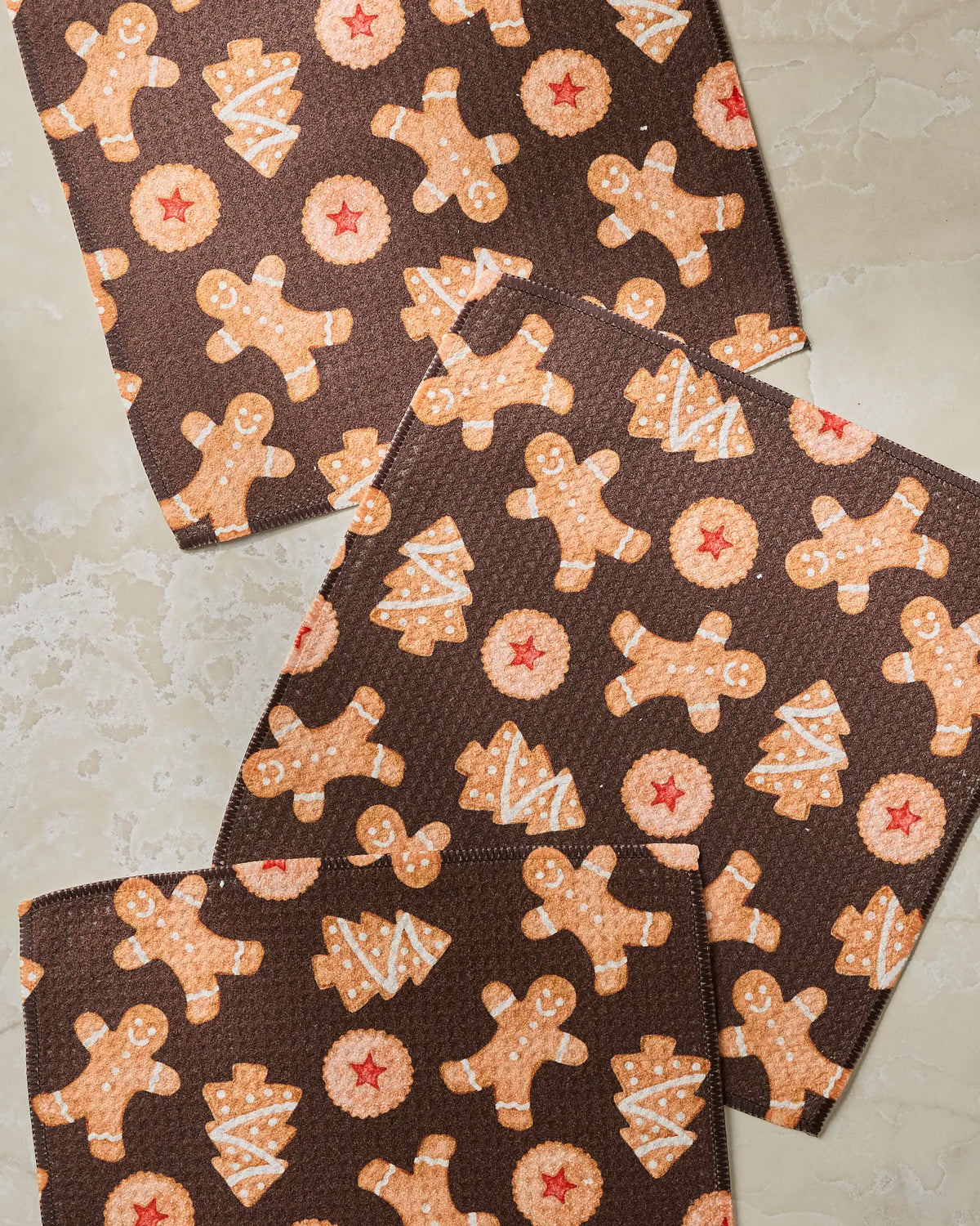 The Geometry “Holiday Cookies” dishcloth set includes three brown cloths adorned with festive patterns of gingerbread men, Christmas trees, and star-shaped cookies on a light marble surface, all crafted from recycled materials for an eco-friendly touch.