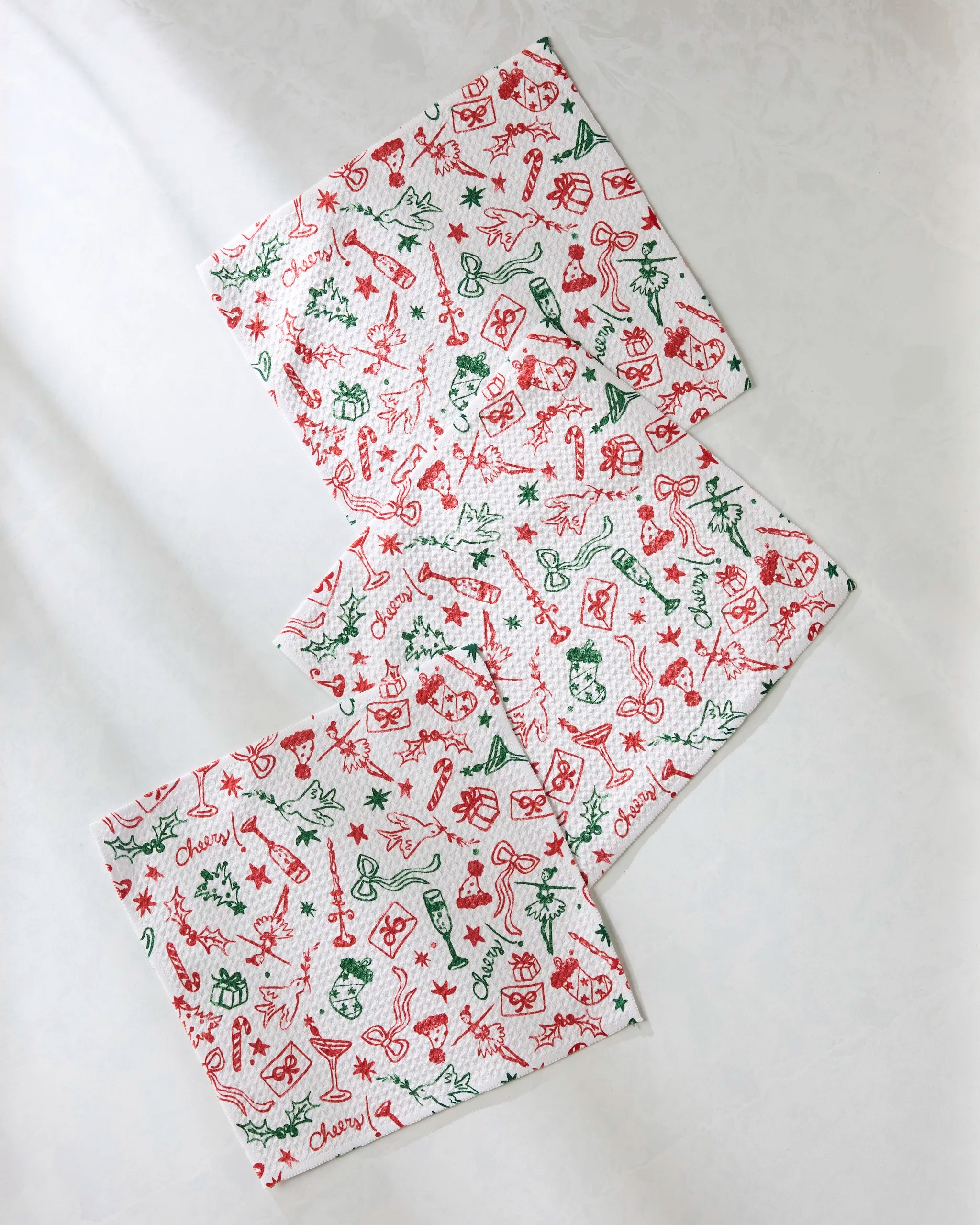 Three white Holiday Doodles Multi dishcloths by Geometry are arranged diagonally on a light surface, each featuring festive red and green holiday patterns with gifts, stars, candy canes, ornaments, and the word "cheery.