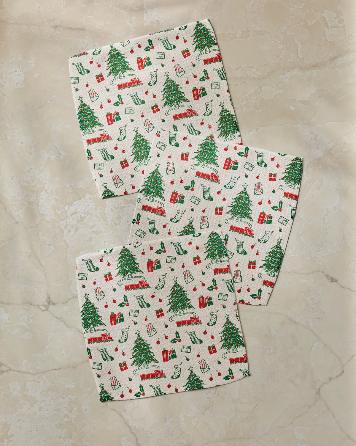 Three super-absorbent Geometry Holiday Jubilee dishtowels, featuring Christmas-themed prints of trees, gifts, stockings, and candy canes, are arranged diagonally on a beige marble surface.