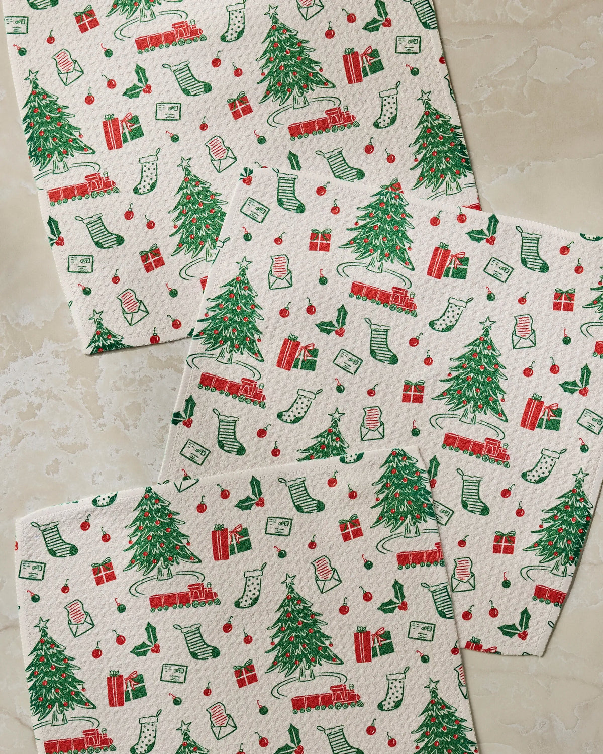 Three festive napkins from Geometry’s Holiday Jubilee super-absorbent dishcloth set, showcasing vibrant red and green Christmas trees, stockings, presents, ornaments, trains, and candy cane designs, are arranged on a beige marble surface.
