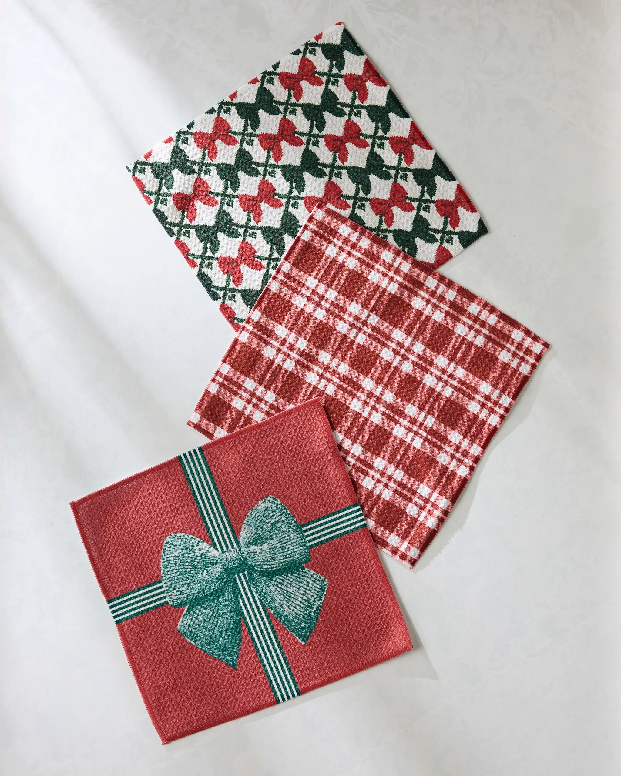 Three Hollis napkins by Geometry, made from recycled materials: one with red and green bows, one with red and white plaid, and one red napkin with a green bow, all displayed on a light surface.