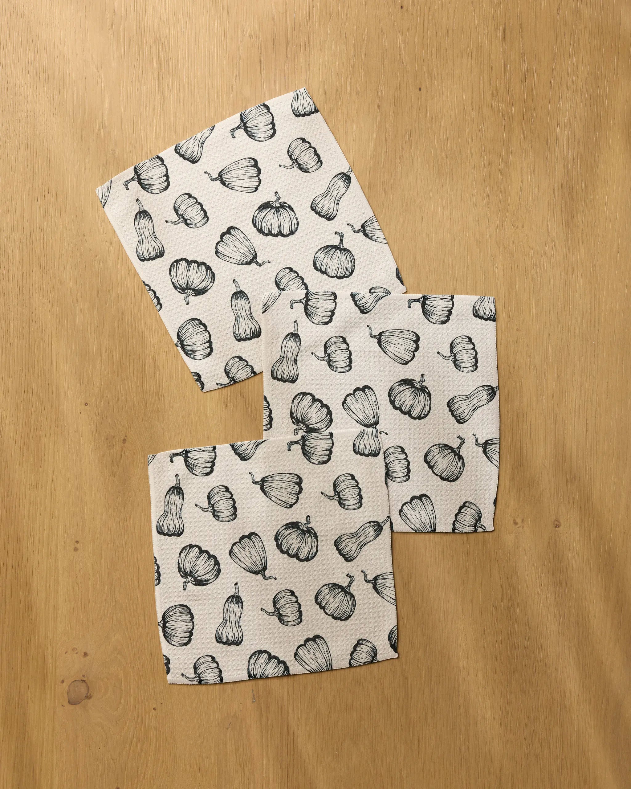 Three Hollow Season dishcloths from Geometry, featuring black line drawings of squash and gourds on white fabric, are arranged on a light wooden surface—combining absorbency and charm for your kitchen.